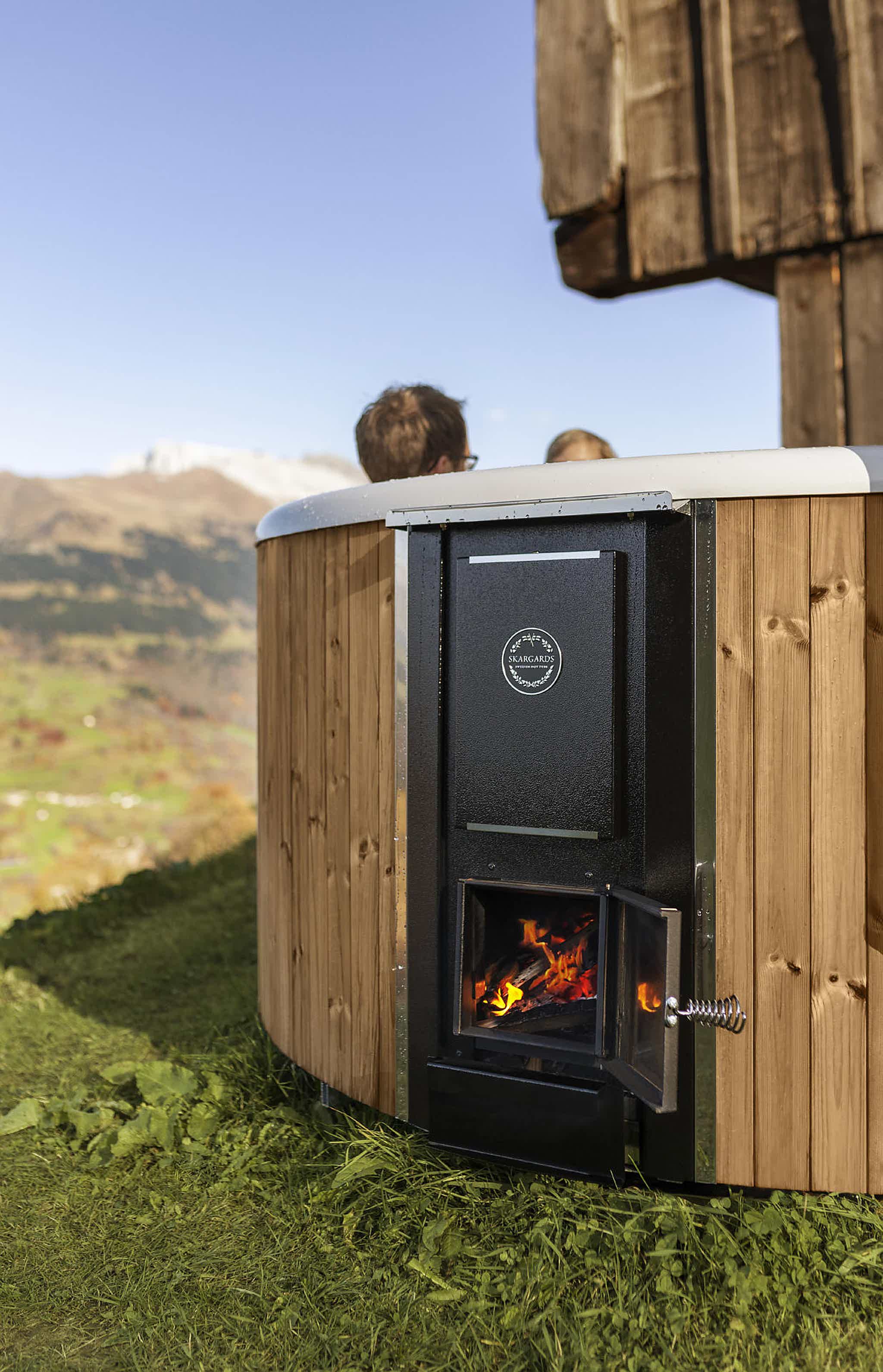 The DualBurn stove of a Skargards Regal 190 hot tub with a couple enjoying a bath, Swiss Alps in the background.