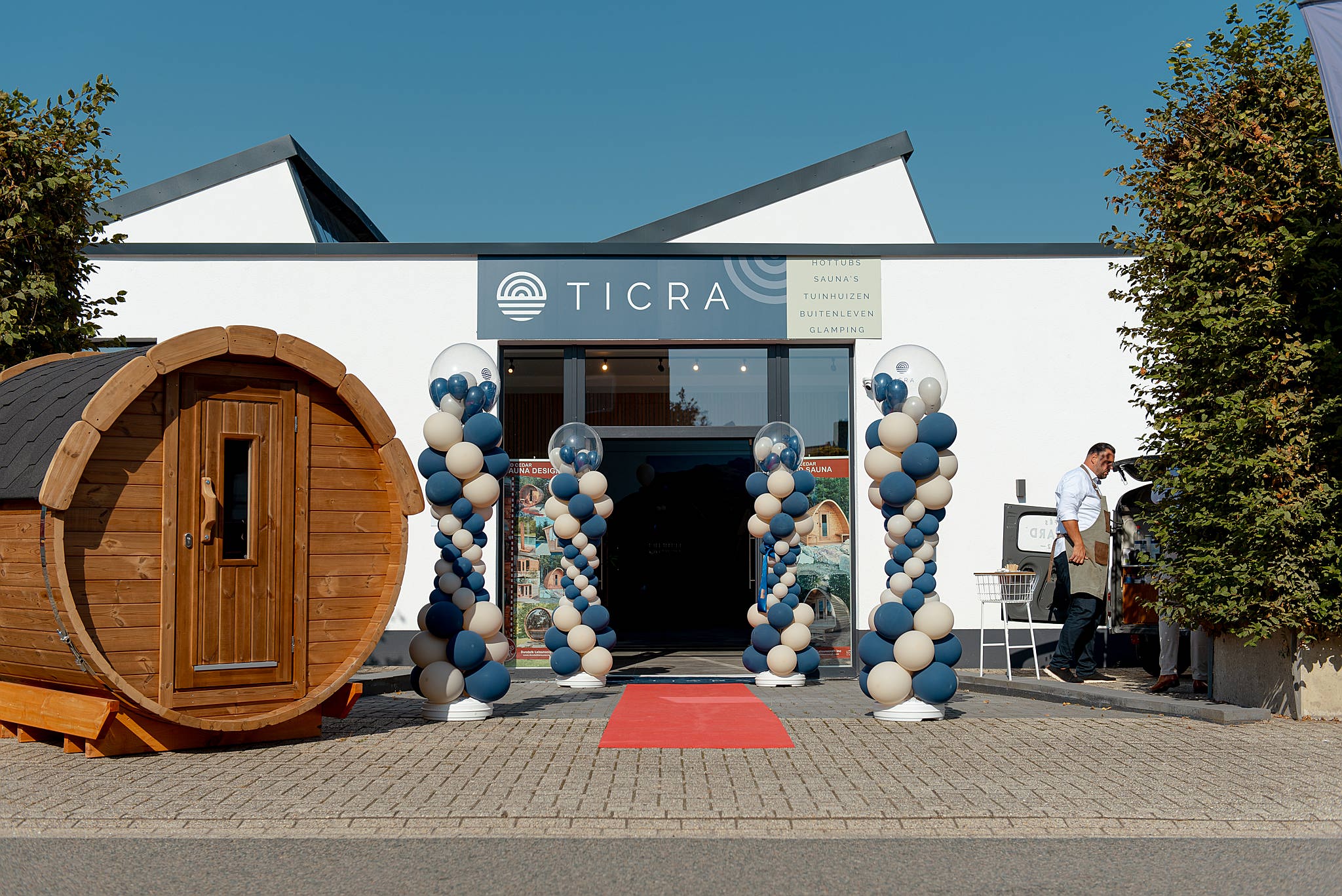 Building with white facade, balloons, wooden barrel, red carpet, person on bench.