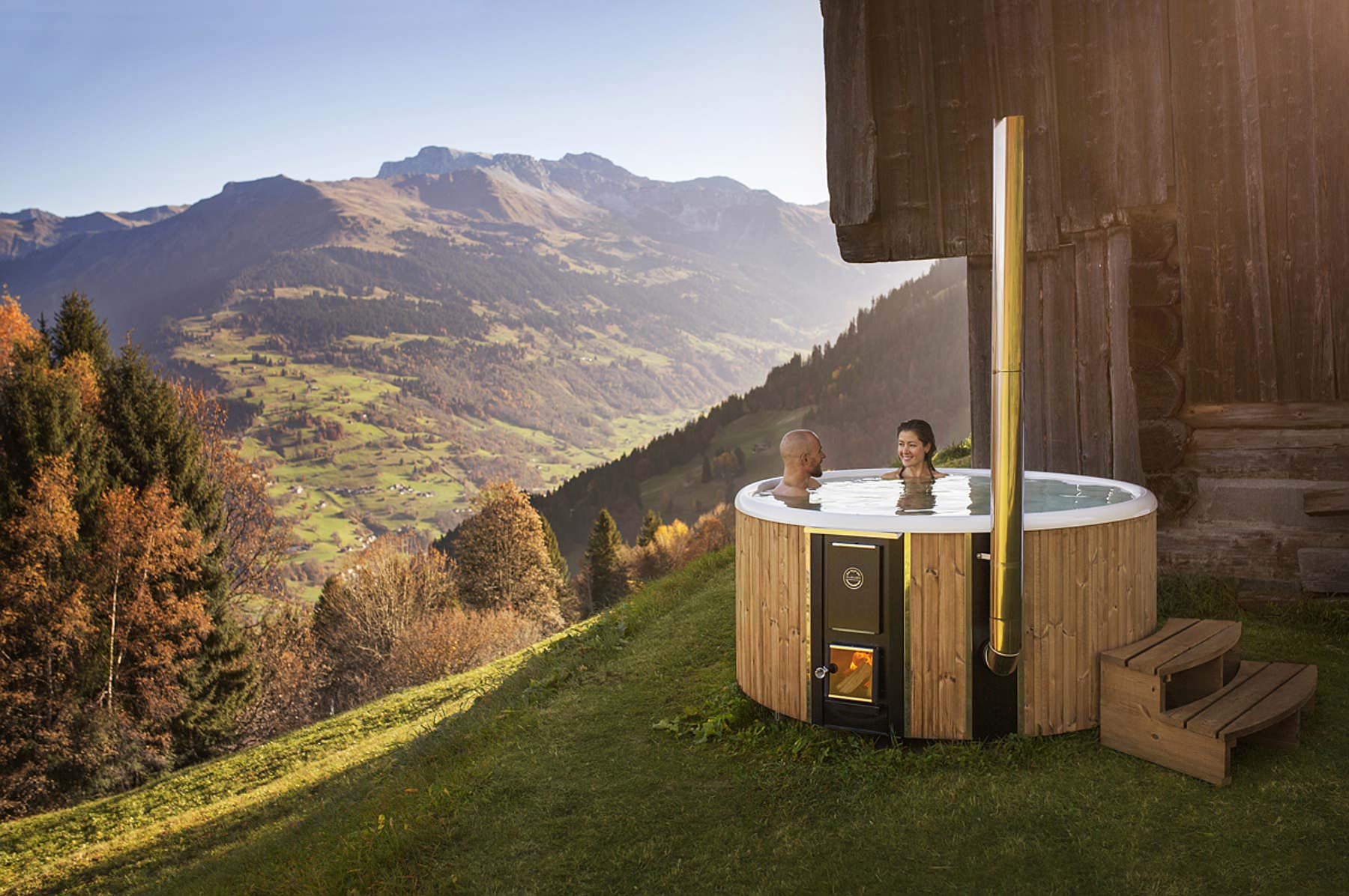 A couple in a wood-fired Skargards hot tub on a hill, enjoying the view of the alps and clear sky.