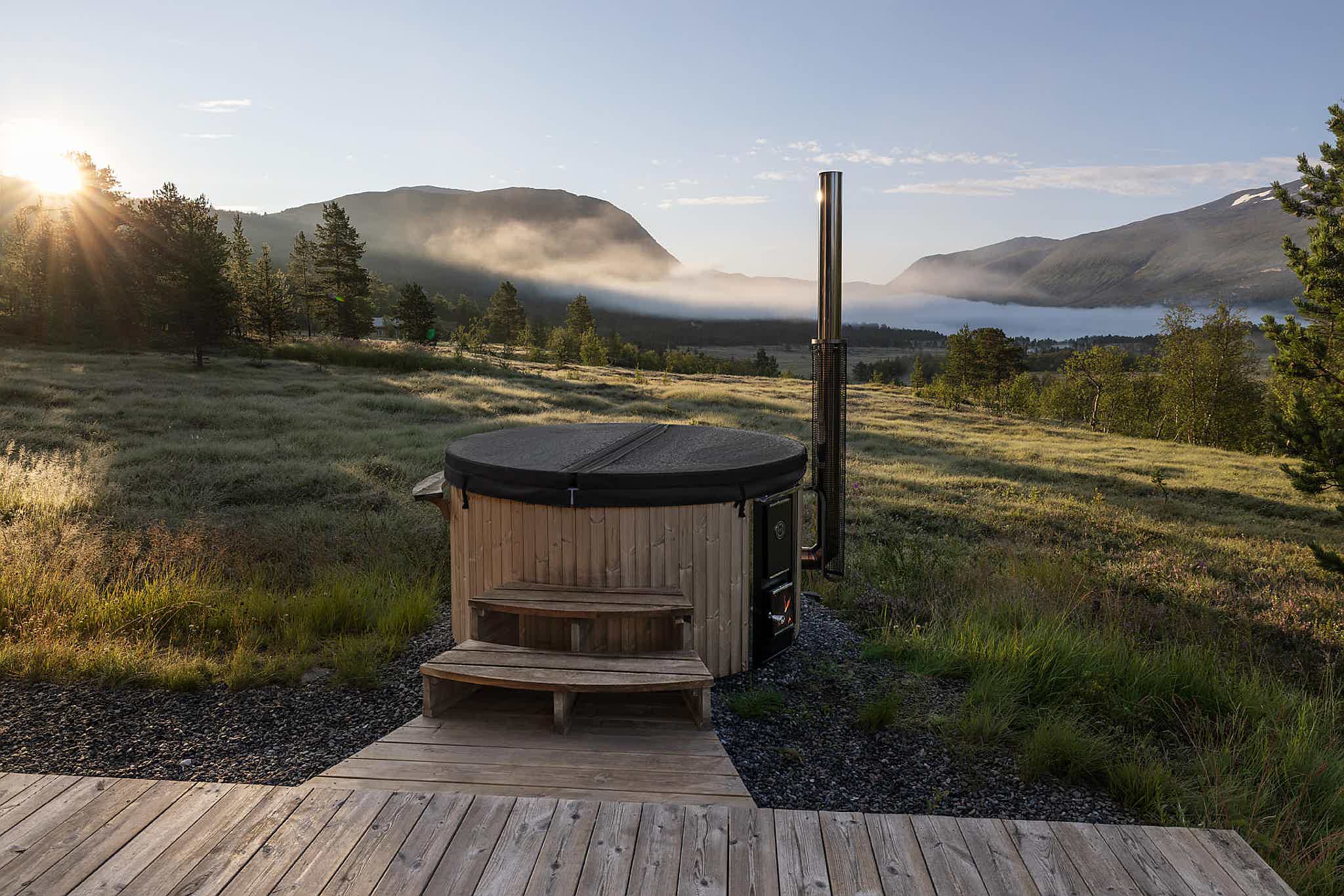 A wood-fired Skargards hot tub with an insulated lid in front of a deck with mountain views and lush greenery, bathed in warm sunlight.