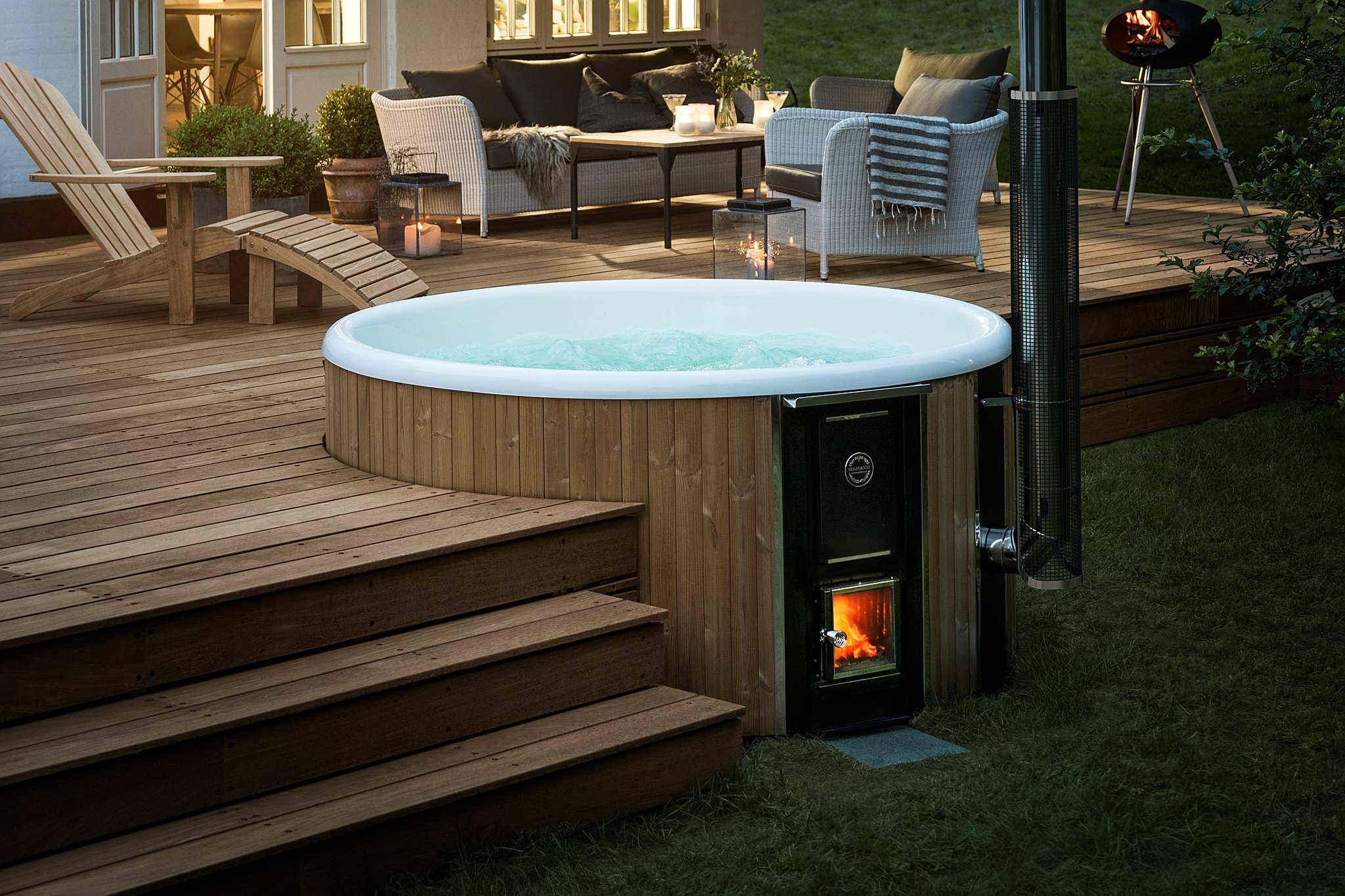 Luxurious backyard with a large round hot tub on a wooden deck, fire pit, and ambient lighting.