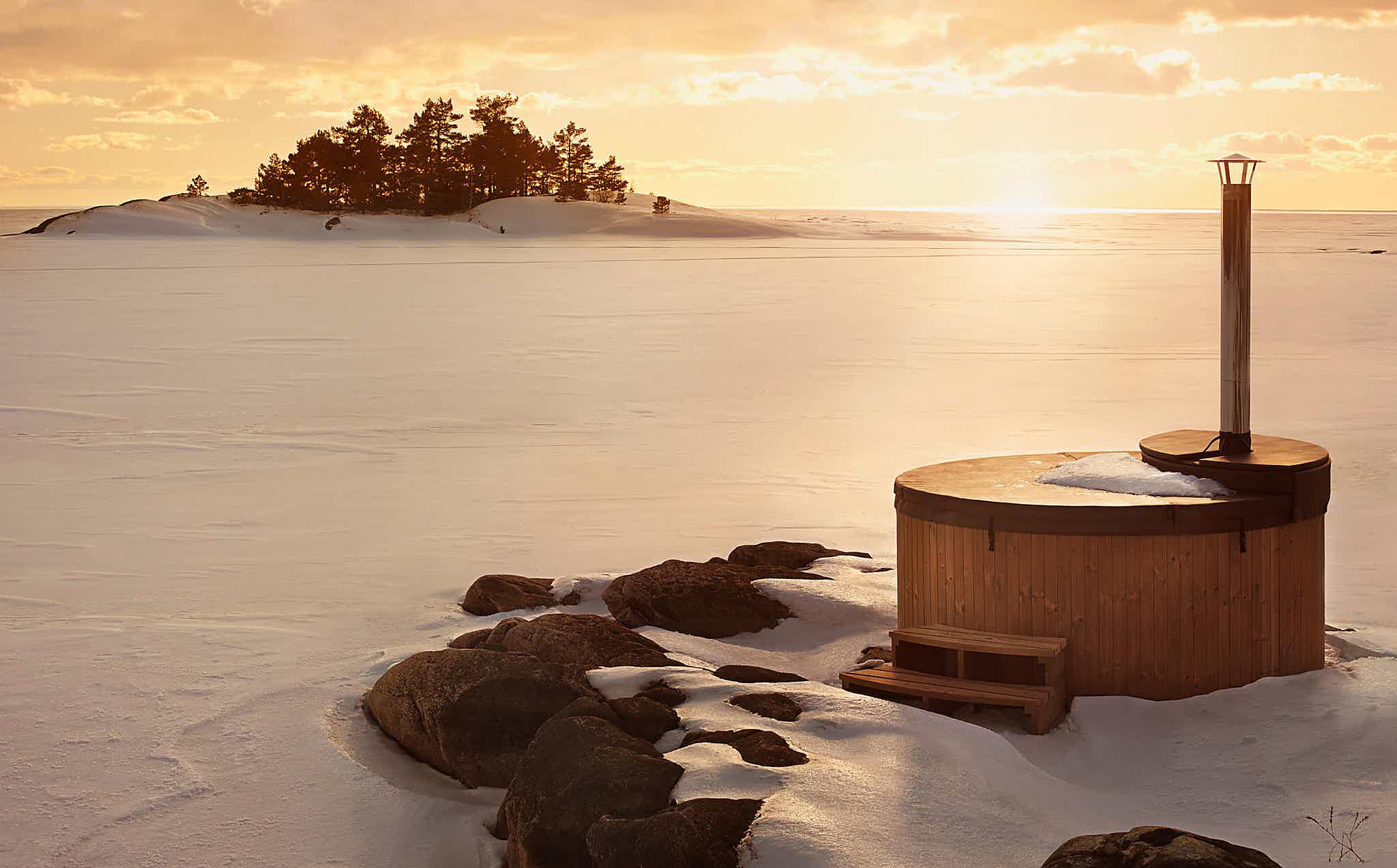 A wooden hot tub on a snowy rocky outcrop with a sunset over mountains.