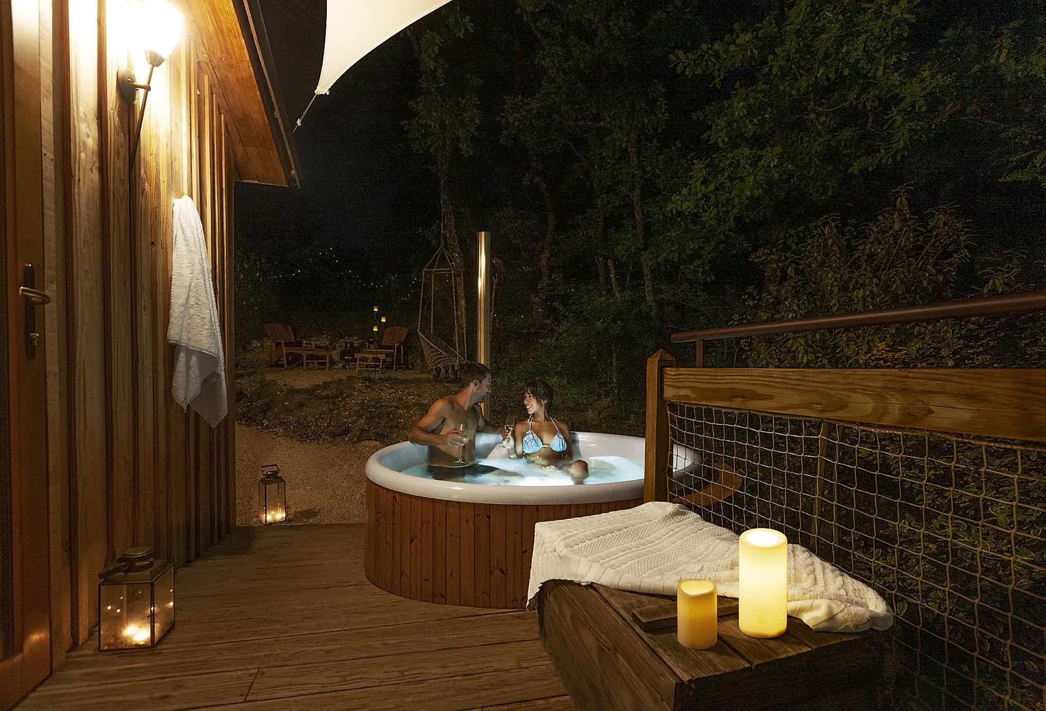 A couple enjoys a Skargards hot tub on a wooden deck, surrounded by trees. A table with lit candles is placed close by.