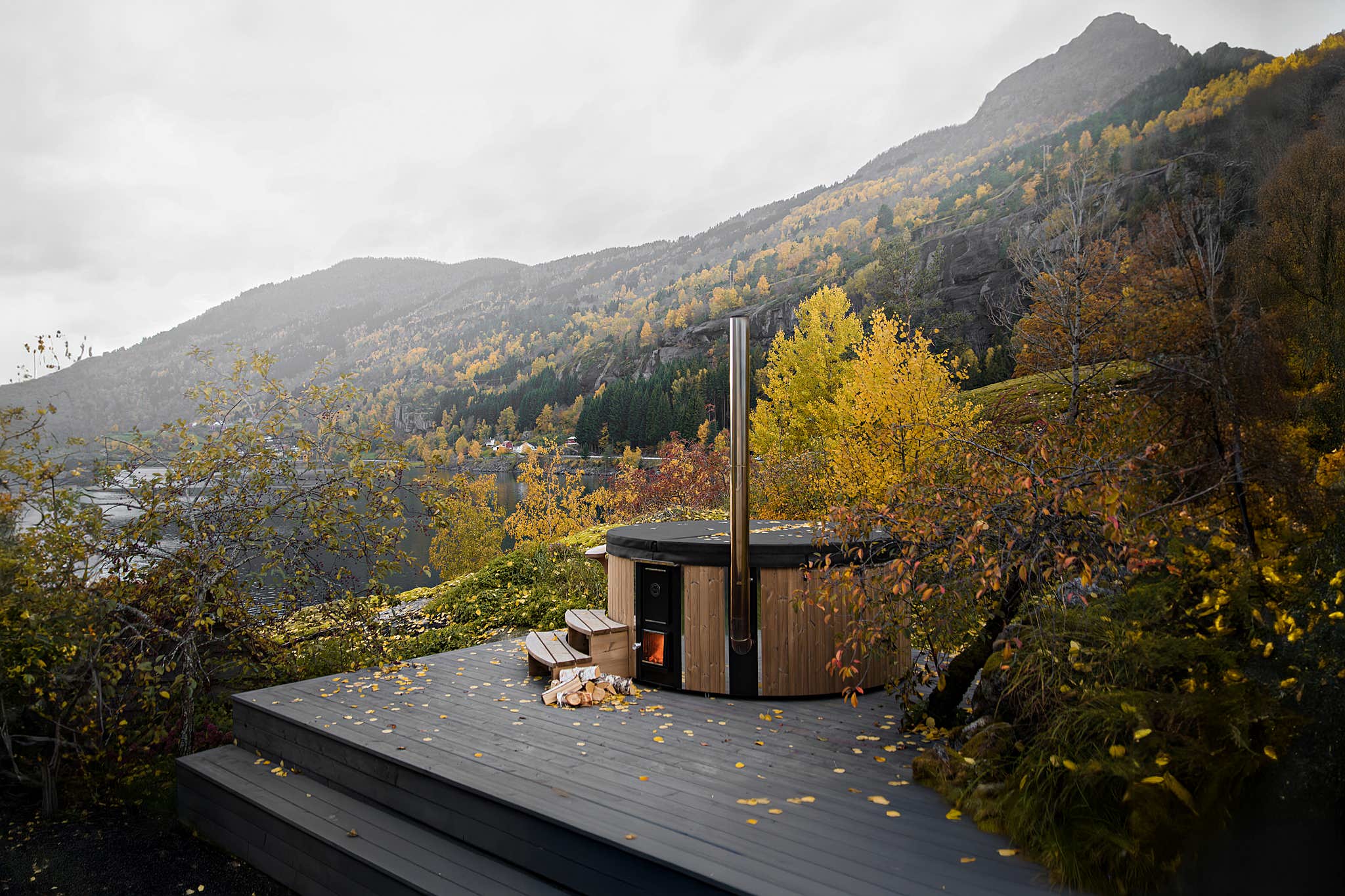 A modern wooden cabin with a chimney on a deck in the mountains, surrounded by trees and a dog on the deck.
