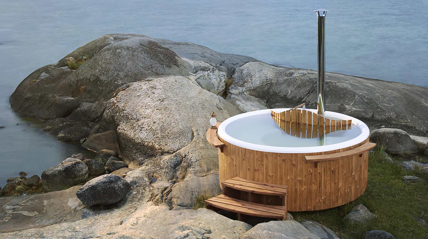 Wooden hot tub on rocky shore, metal pole, serene scene.
