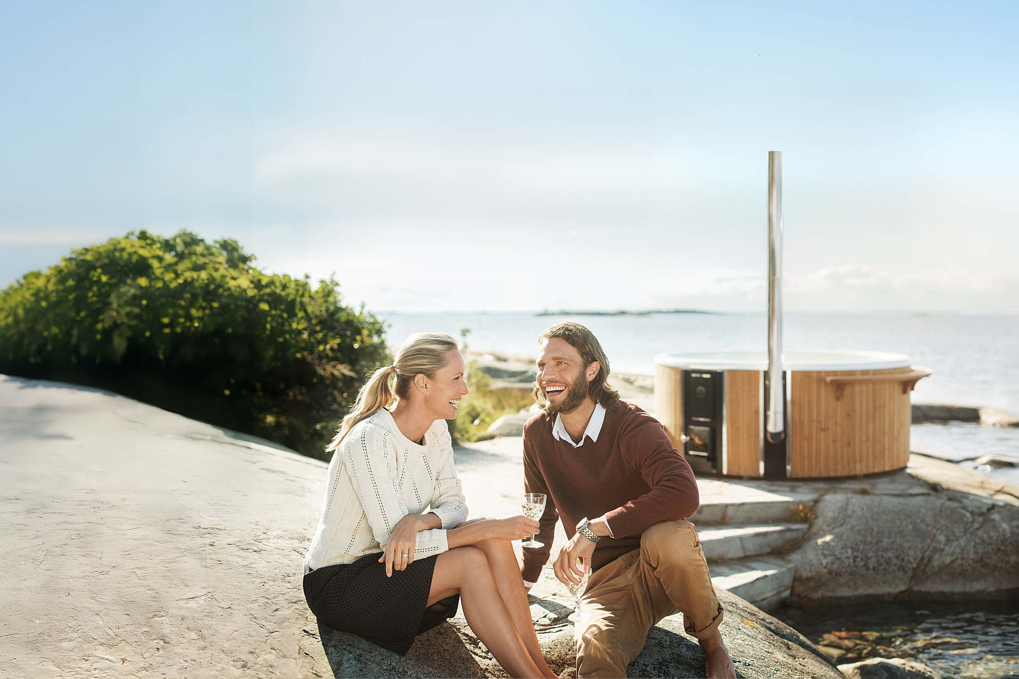 A couple sits by the ocean, smiling, with a wooden structure nearby.