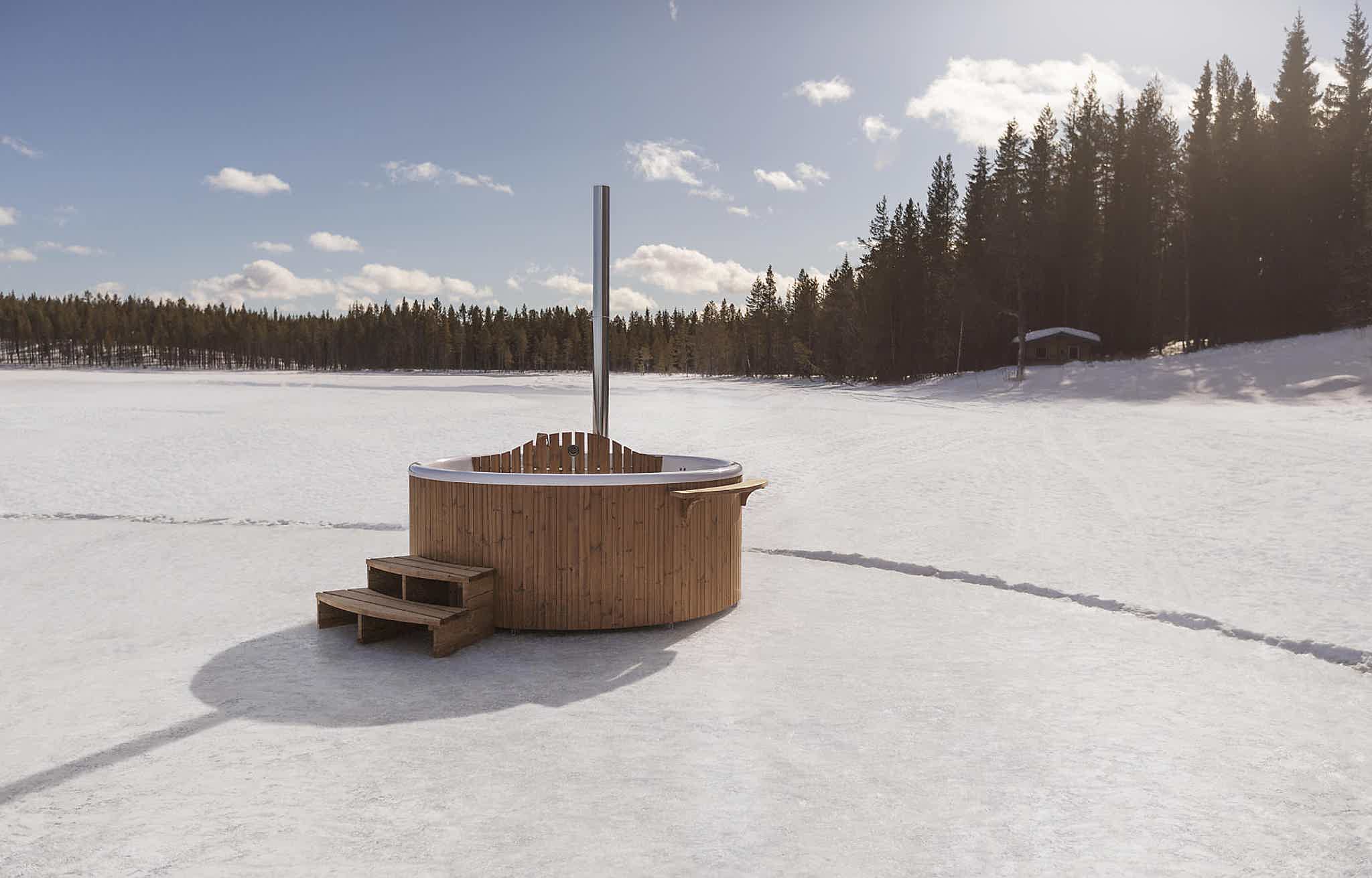 A wooden hot tub on a snowy lake with wooden steps and clear blue sky.