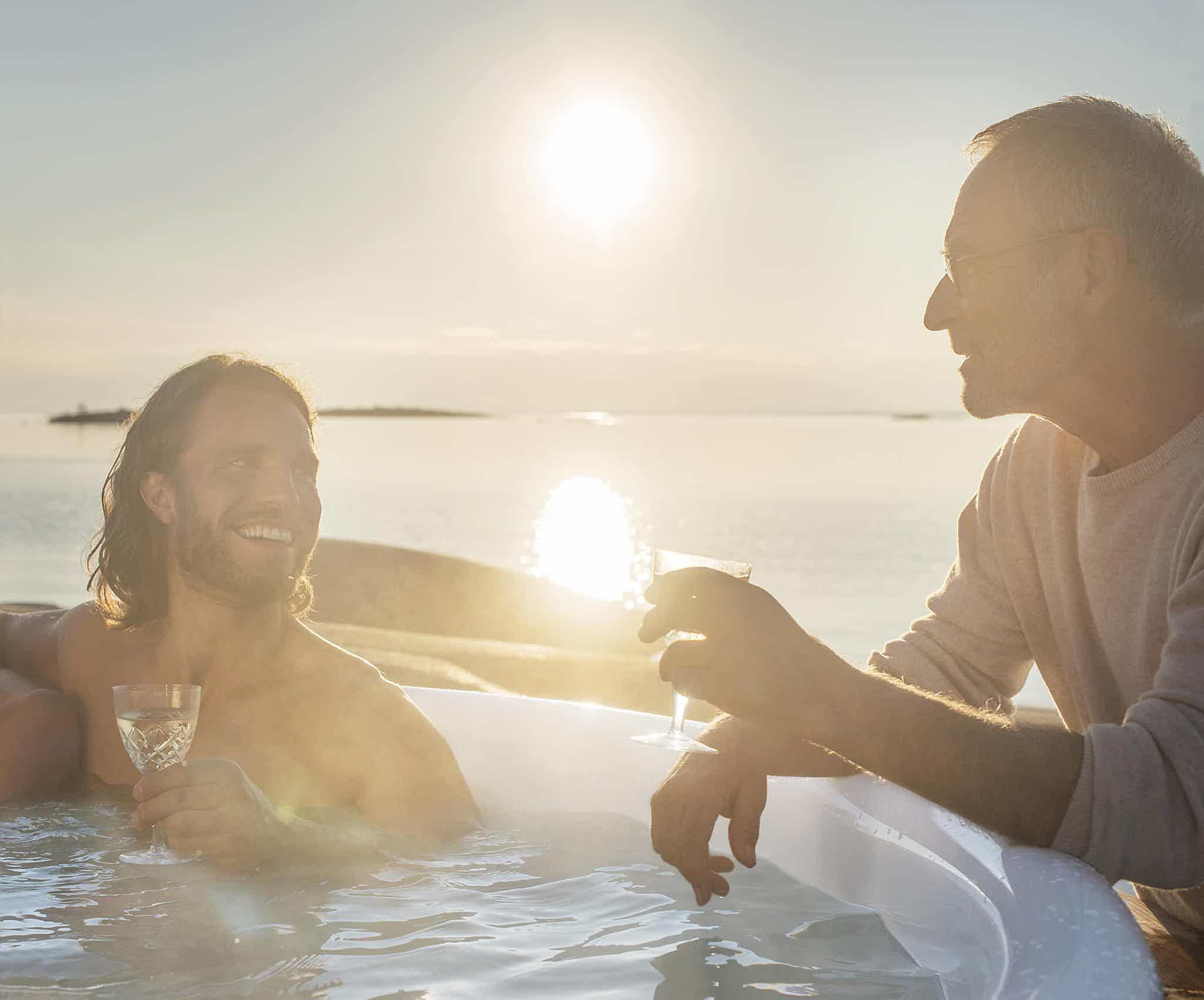 Two men chat at sunset near a Skargards hot tub, one standing outside and the other relaxing in the warm water, both holding drinks.