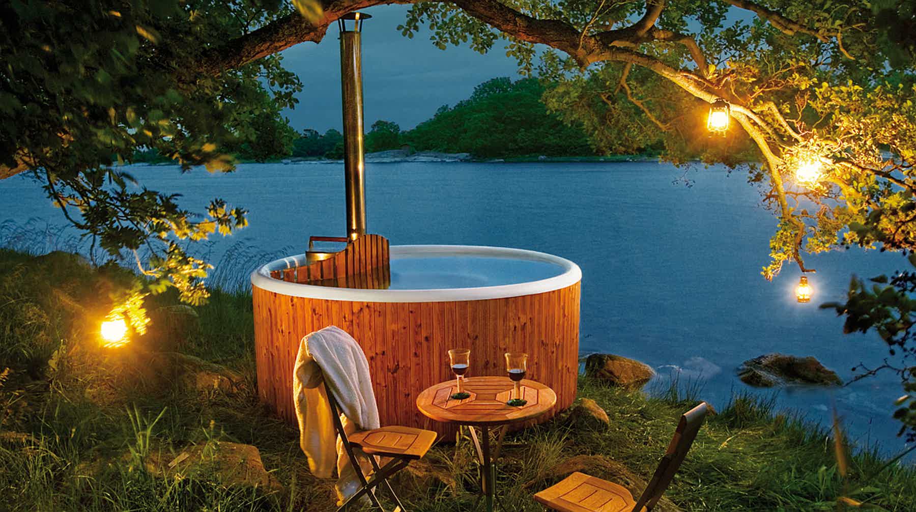 A serene setting with a wooden hot tub, chair, and table by a lake and mountains, lit warmly.