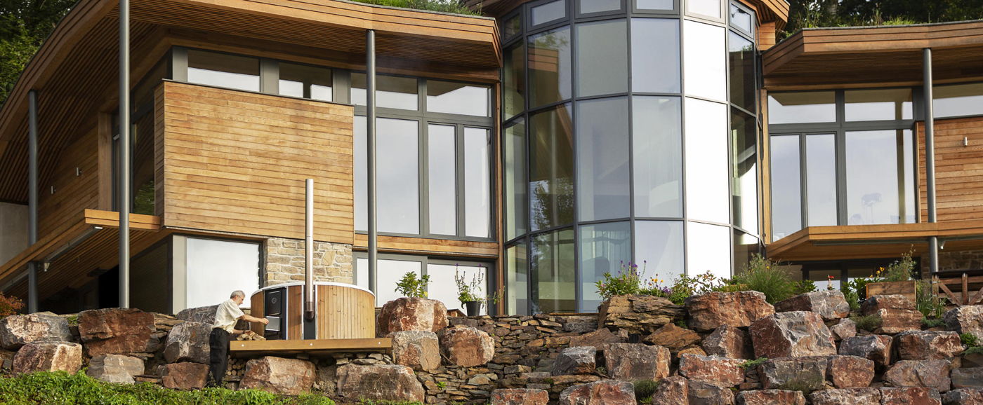 A man filling the stove of the Skargards Regal wood-burning hot tub which is standing in front of a huge sustainable house