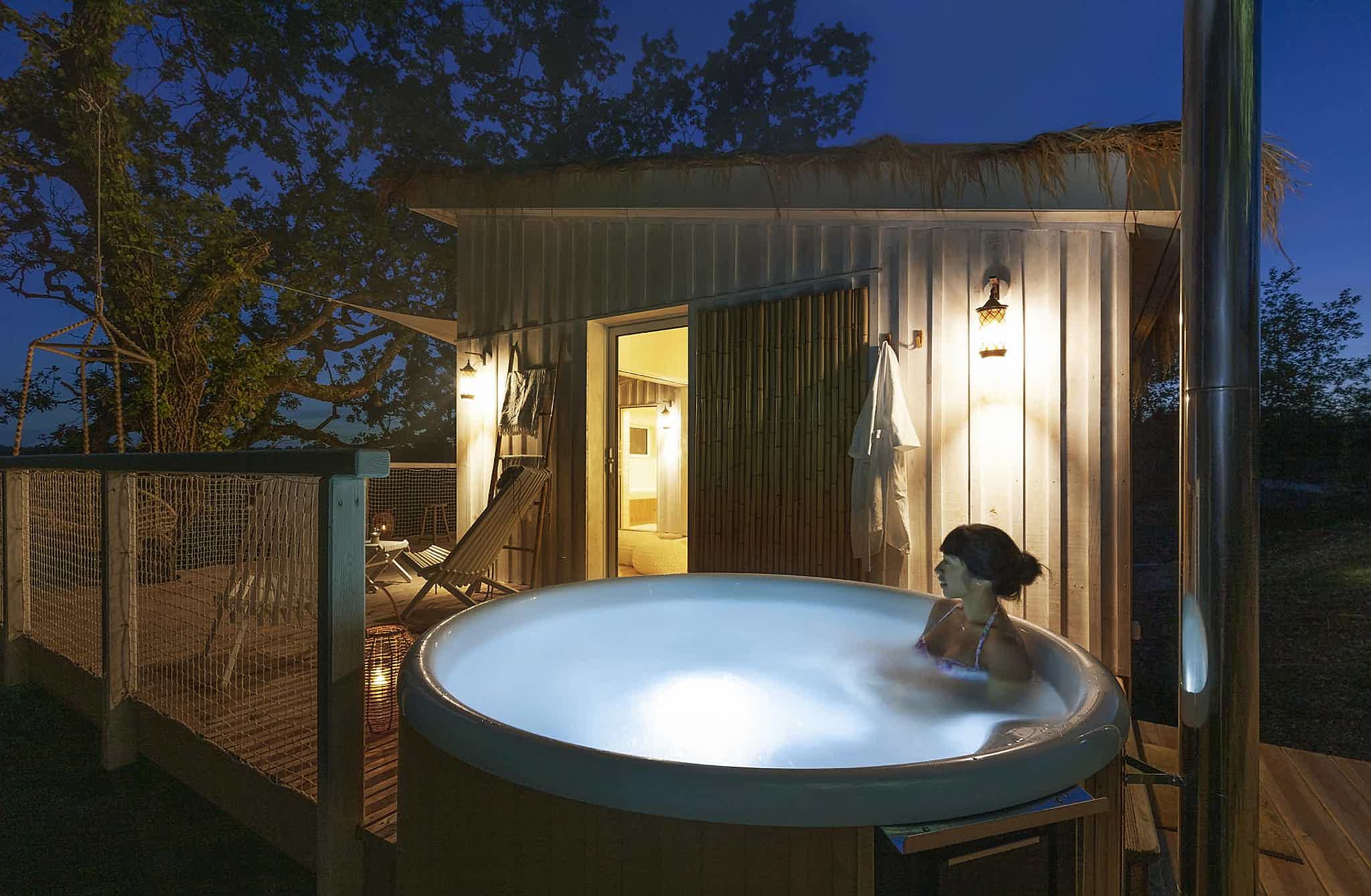 A woman relaxes in a Skargards Regal hot tub on a wooden deck, cozy lights light the scene.