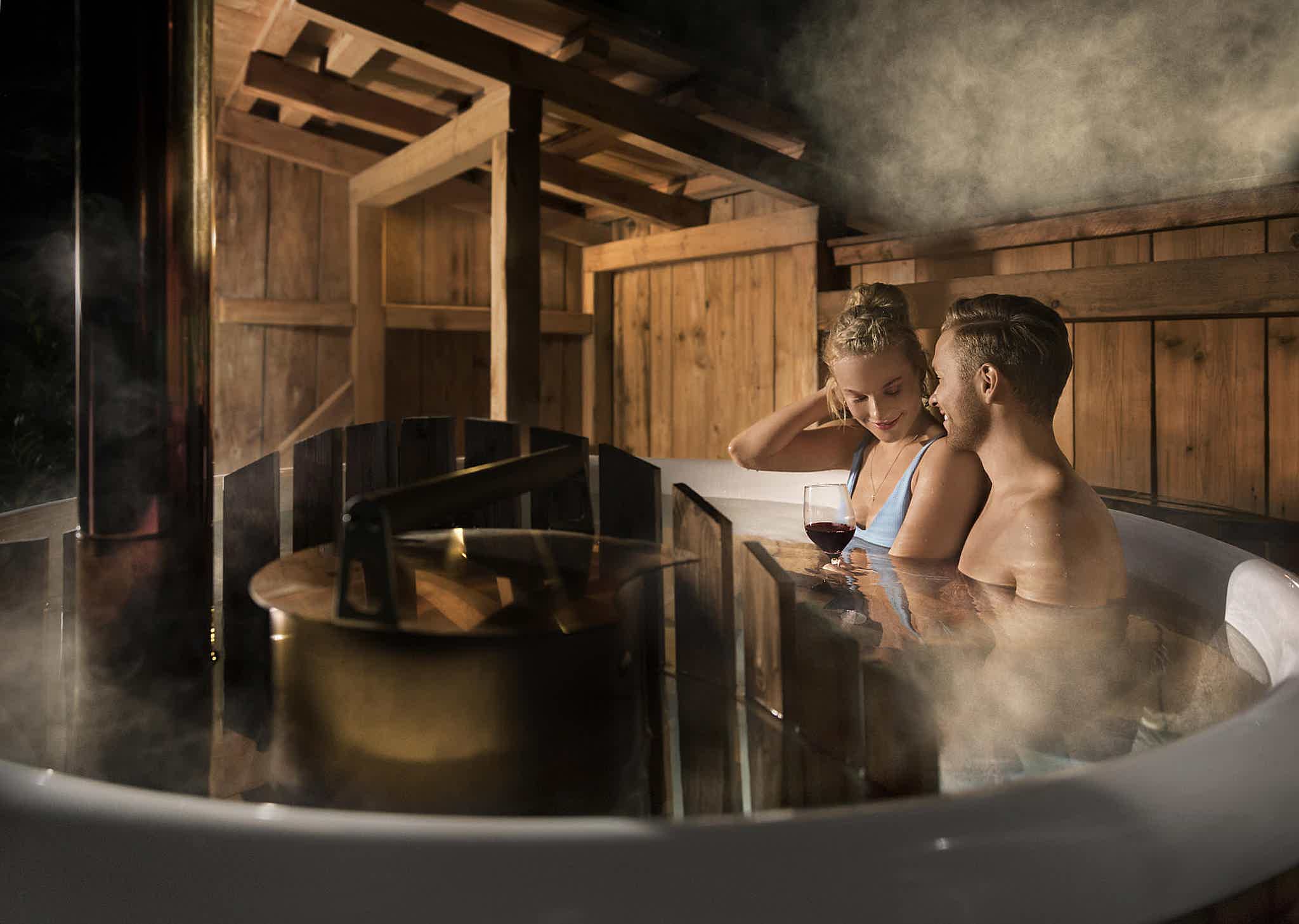 A couple enjoys a drink in a Skargards Panel 190 hot tub, the stove of the hot tub is visible in the foreground.