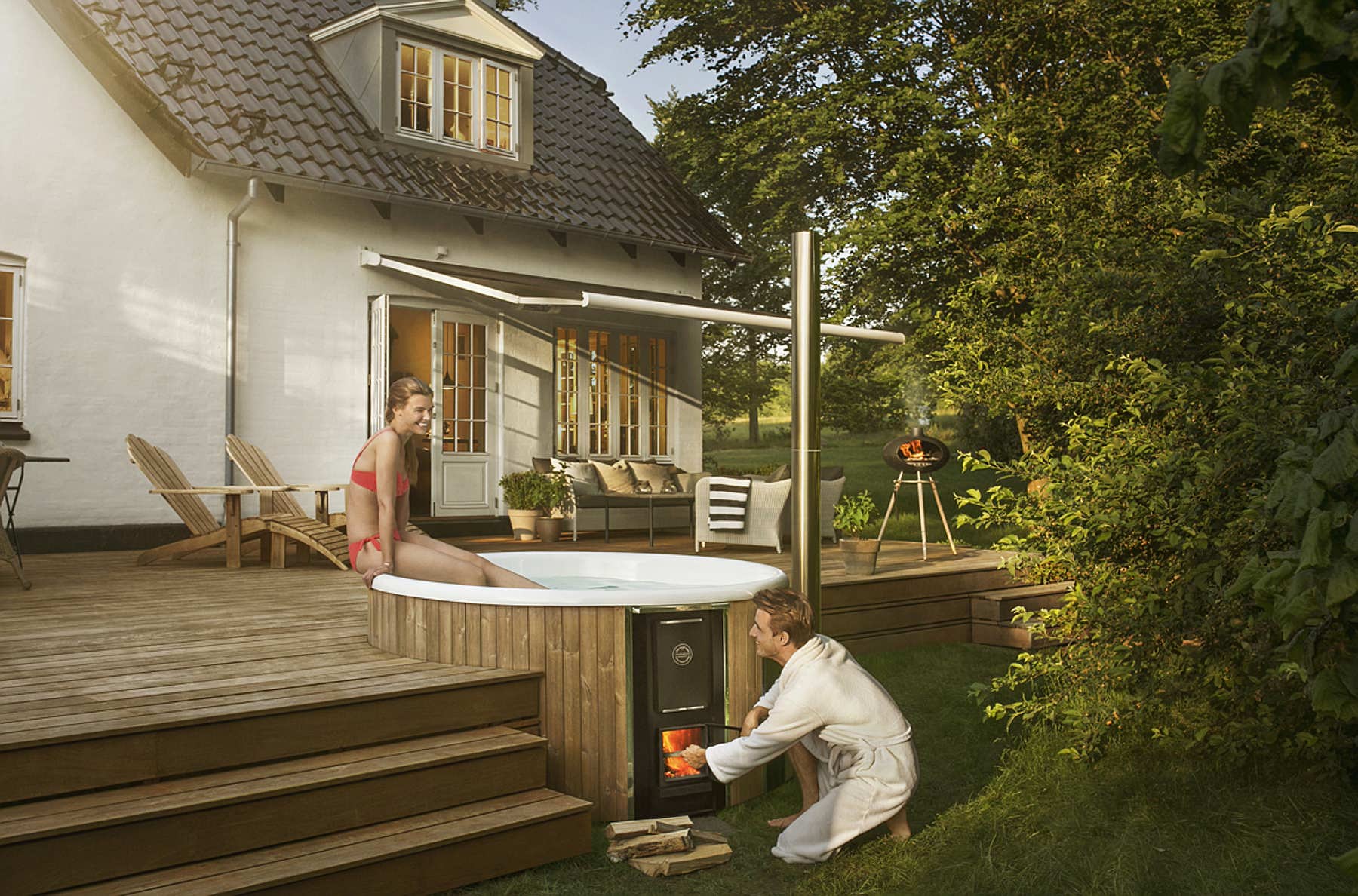 A woman enjoys a wood-fired Skargards hot tub on a wooden terrace while a man heats the stove.