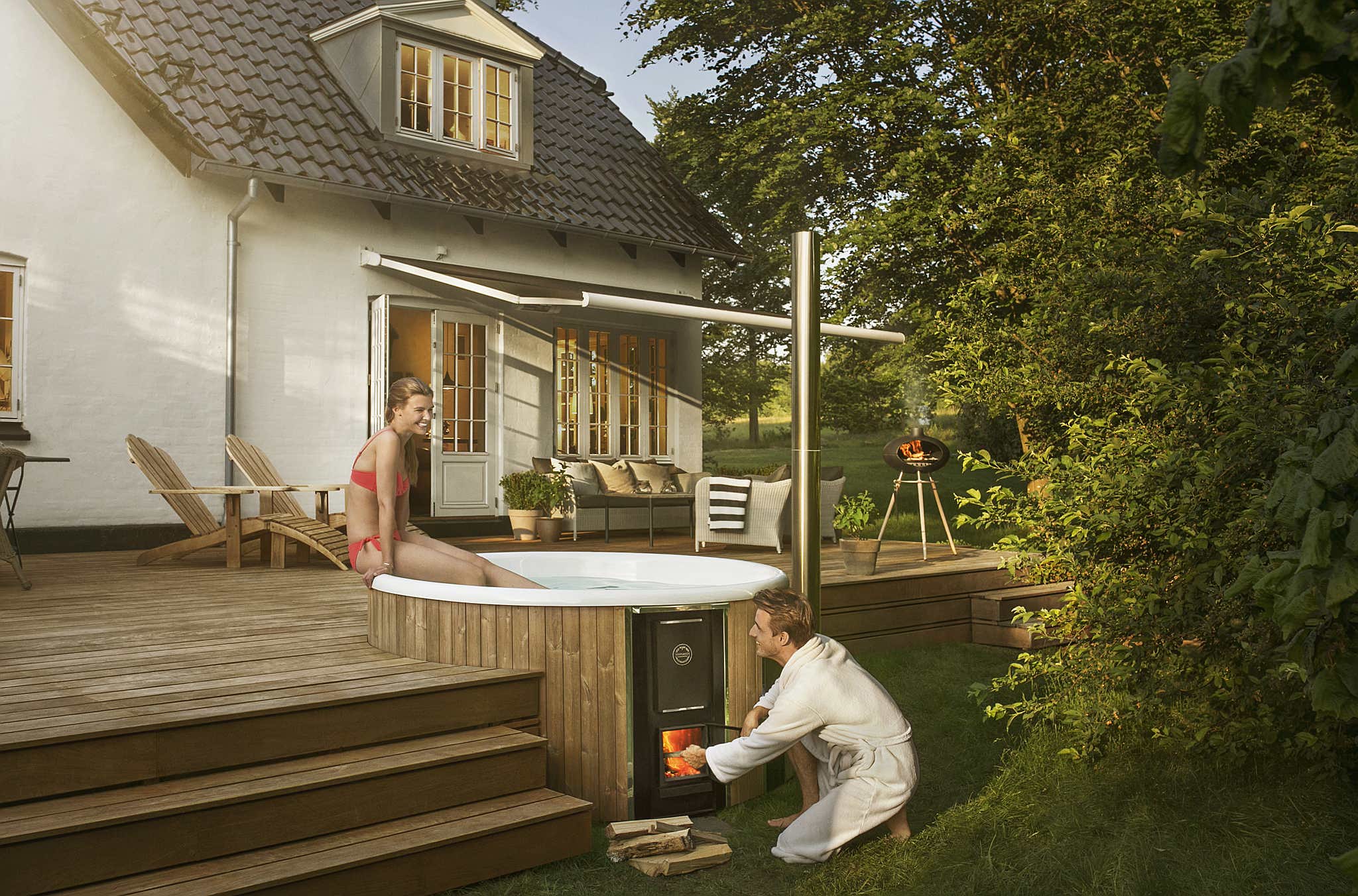 A woman enjoys a wood-fired Skargards hot tub on a wooden terrace while a man heats the stove.