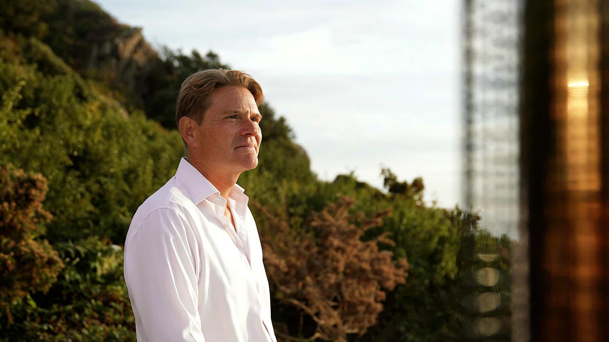 A man in a white shirt stands by his hot tub, watching the sunset. Trees and a hill are in the background.