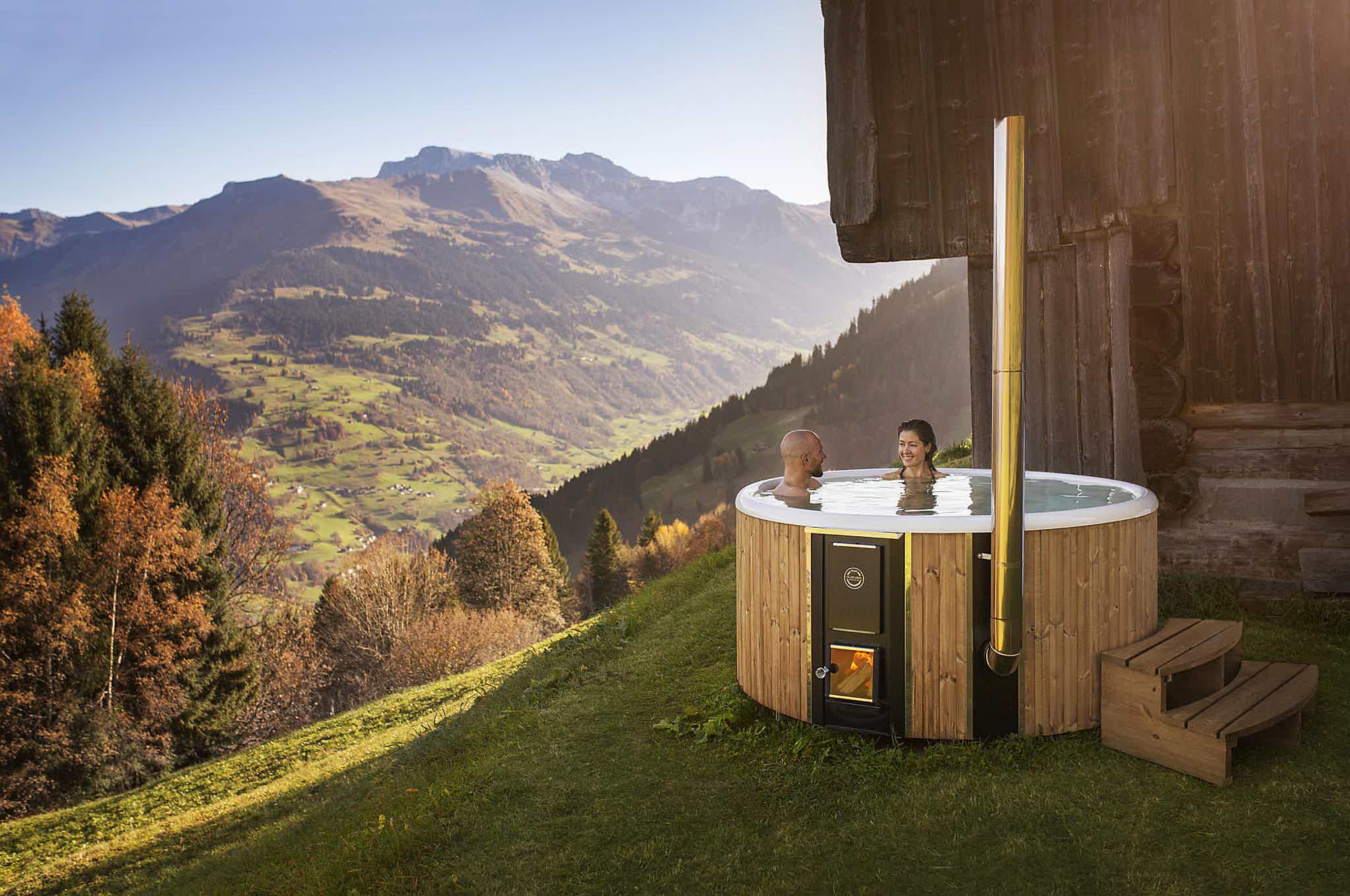 A couple in a wood-fired Skargards hot tub on a hill, enjoying the view of the alps and clear sky.