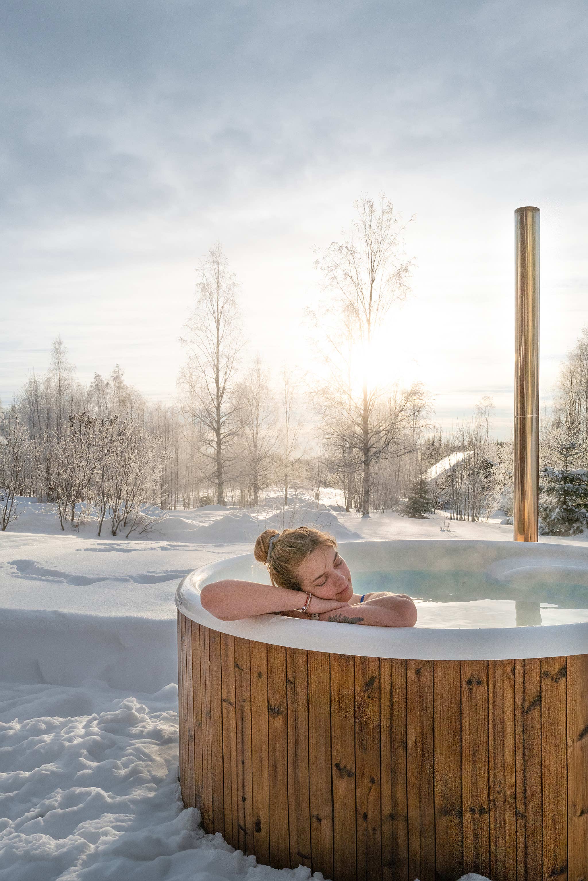 A woman relaxes in a wooden hot tub amidst snow-covered trees.
