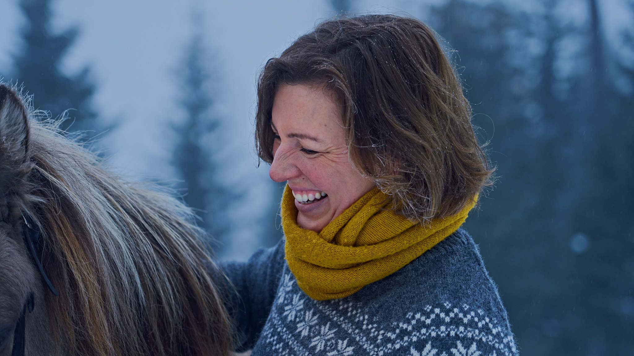 A woman with short brown hair and a yellow scarf smiles while petting a horse in a snowy forest.