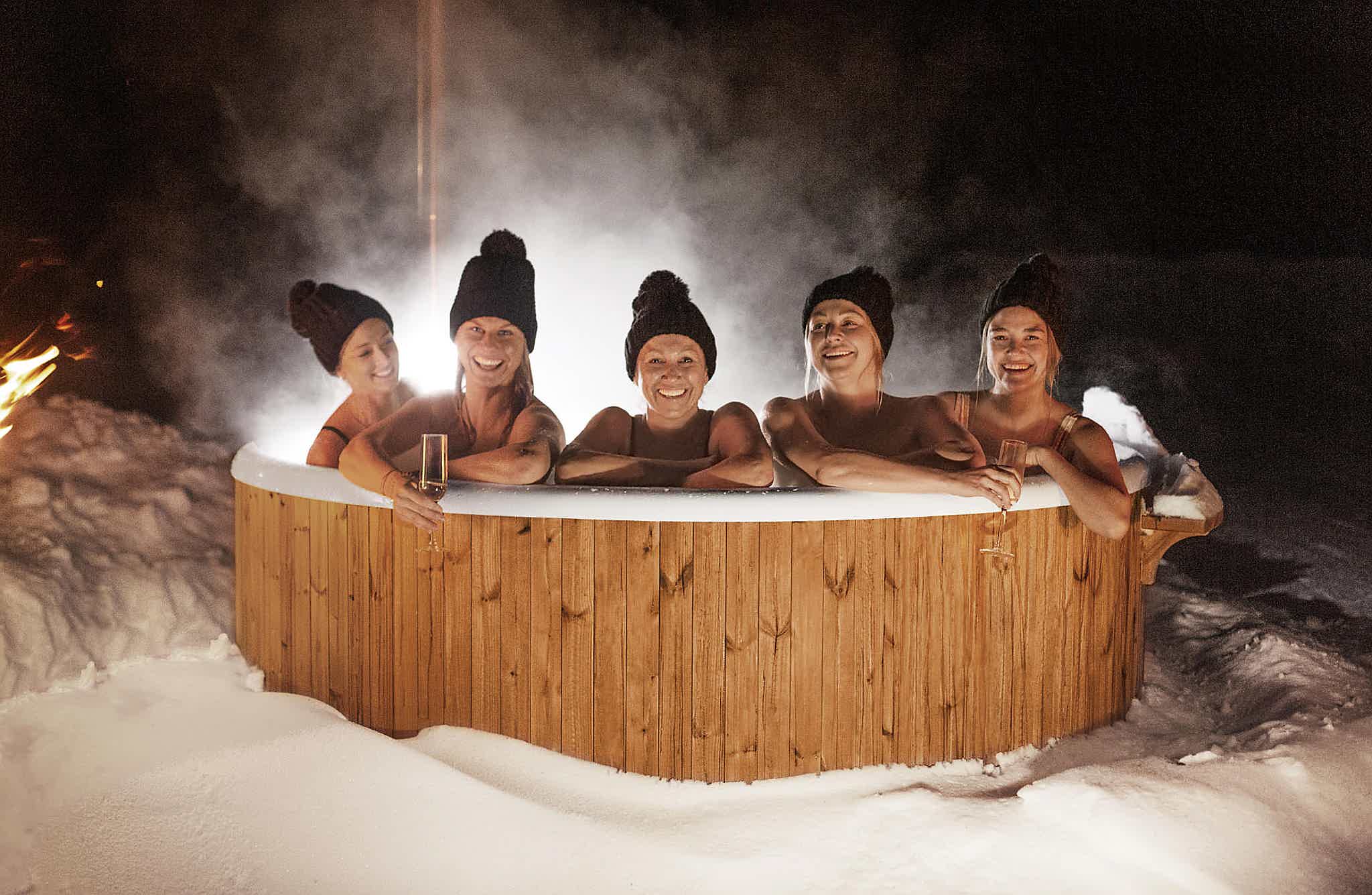 Five people in a steaming wooden Skargards hot tub surrounded by deep snow, wearing black hats, enjoying a warm fire.