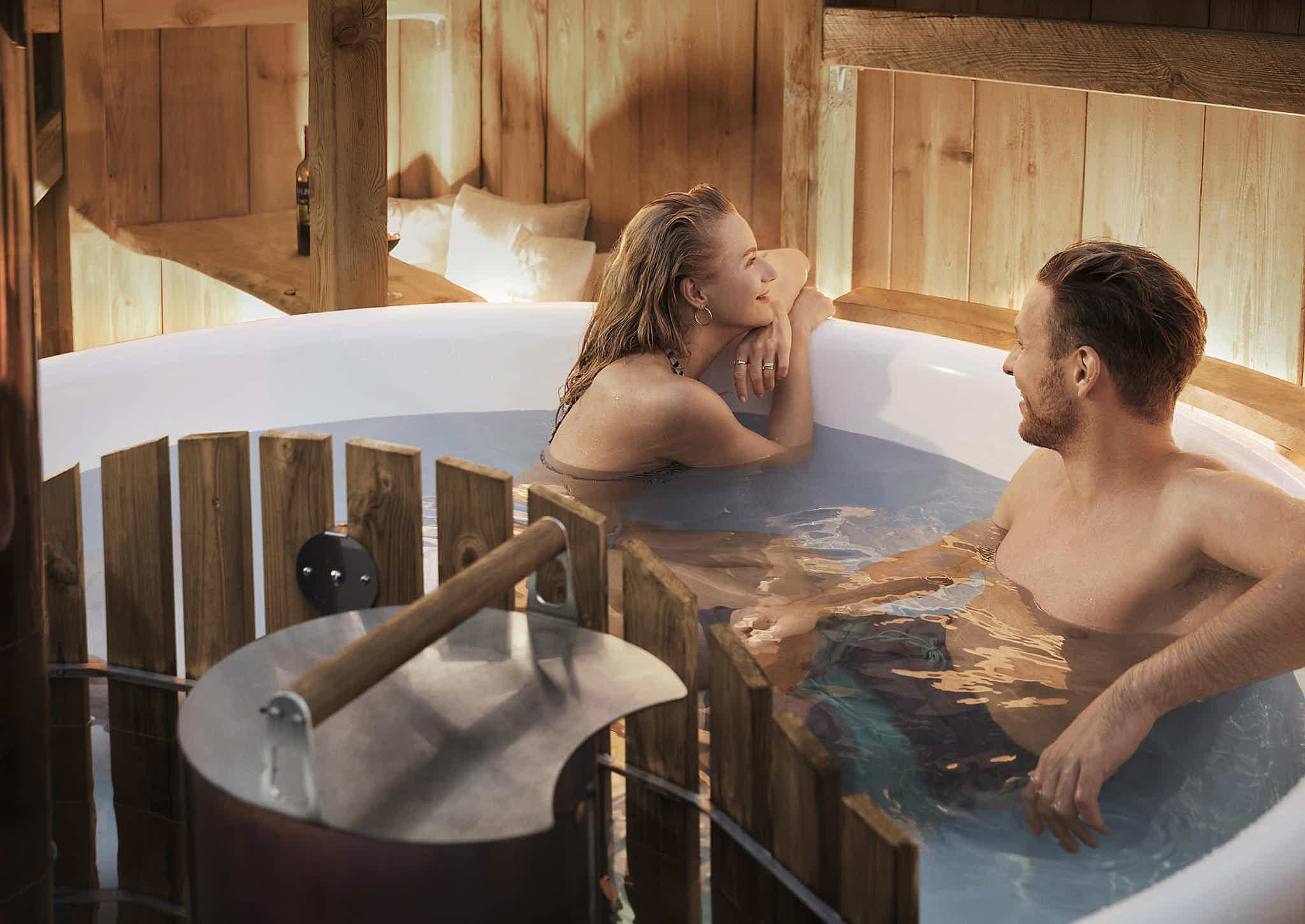 A couple enjoys the warm water of a Skargards Panel 190 hot tub, covered by a wooden shed. The woman faces to the outside and smiles, the man smiles at her.