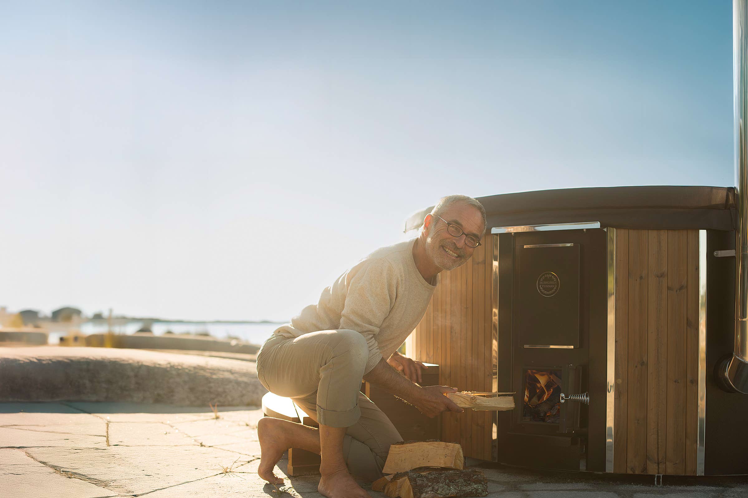 An elderly man is kneeling by a wood stove, smiling at the camera.