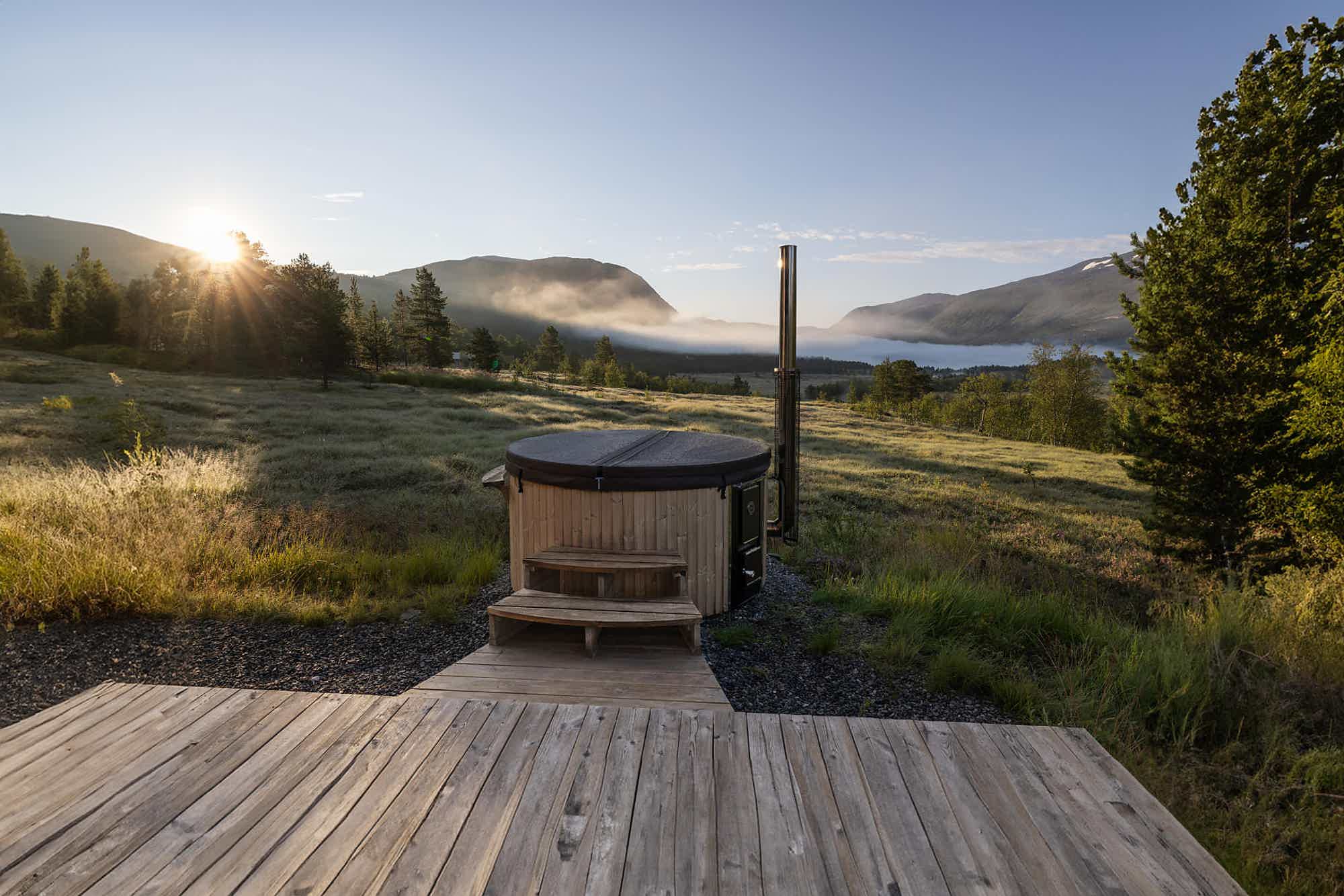 A wood-fired Skargards hot tub with an insulated lid on a wooden deck with mountain views in warm sunlight.