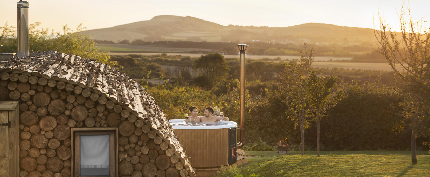 A couple enjoying the wood-burning hot tub Skargards Regal next to a wooden house at a glamping side in United Kingdom