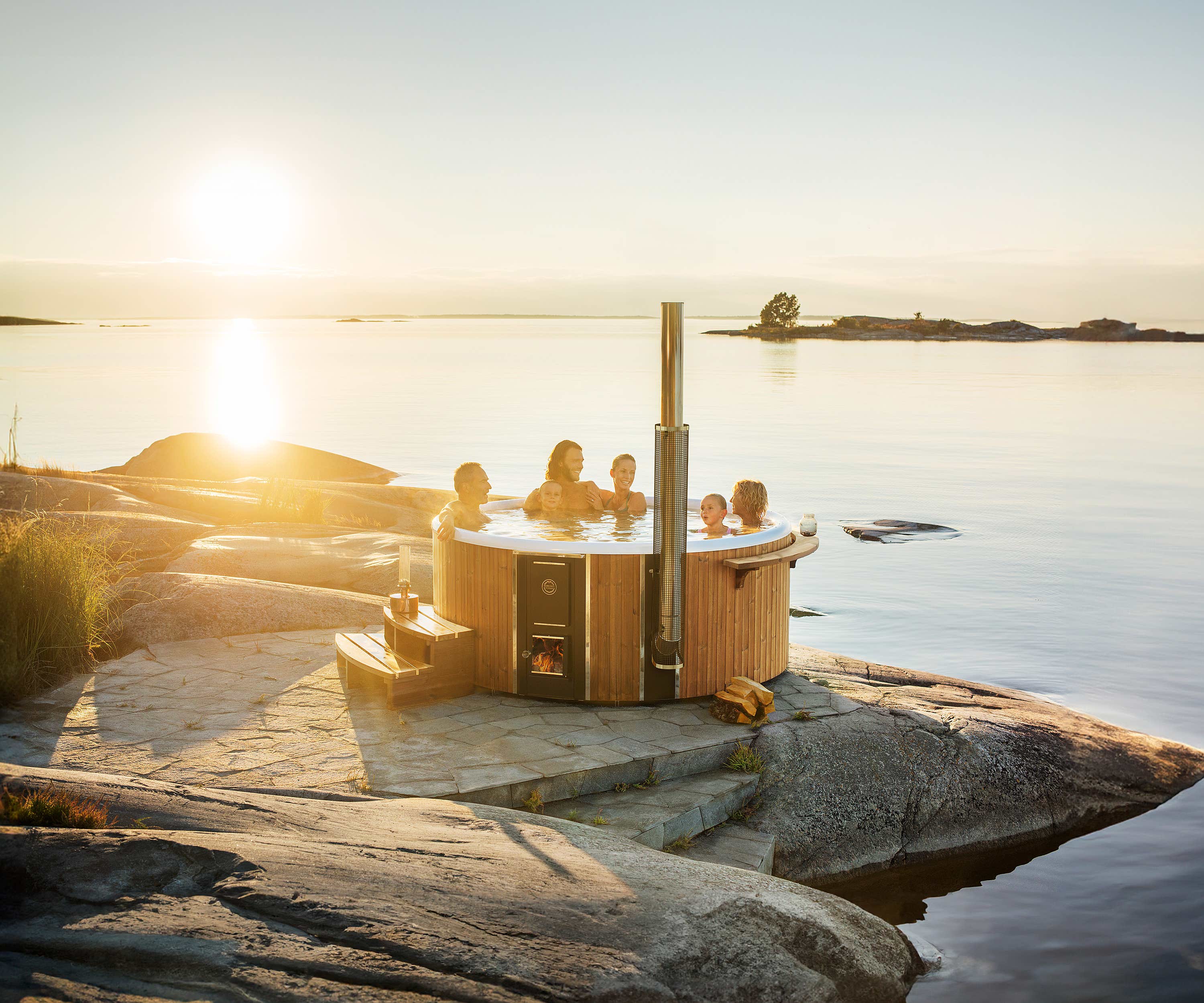 People enjoying a Skargards Regal hot tub by the shore at sunset, surrounded by rocks and a chimney.