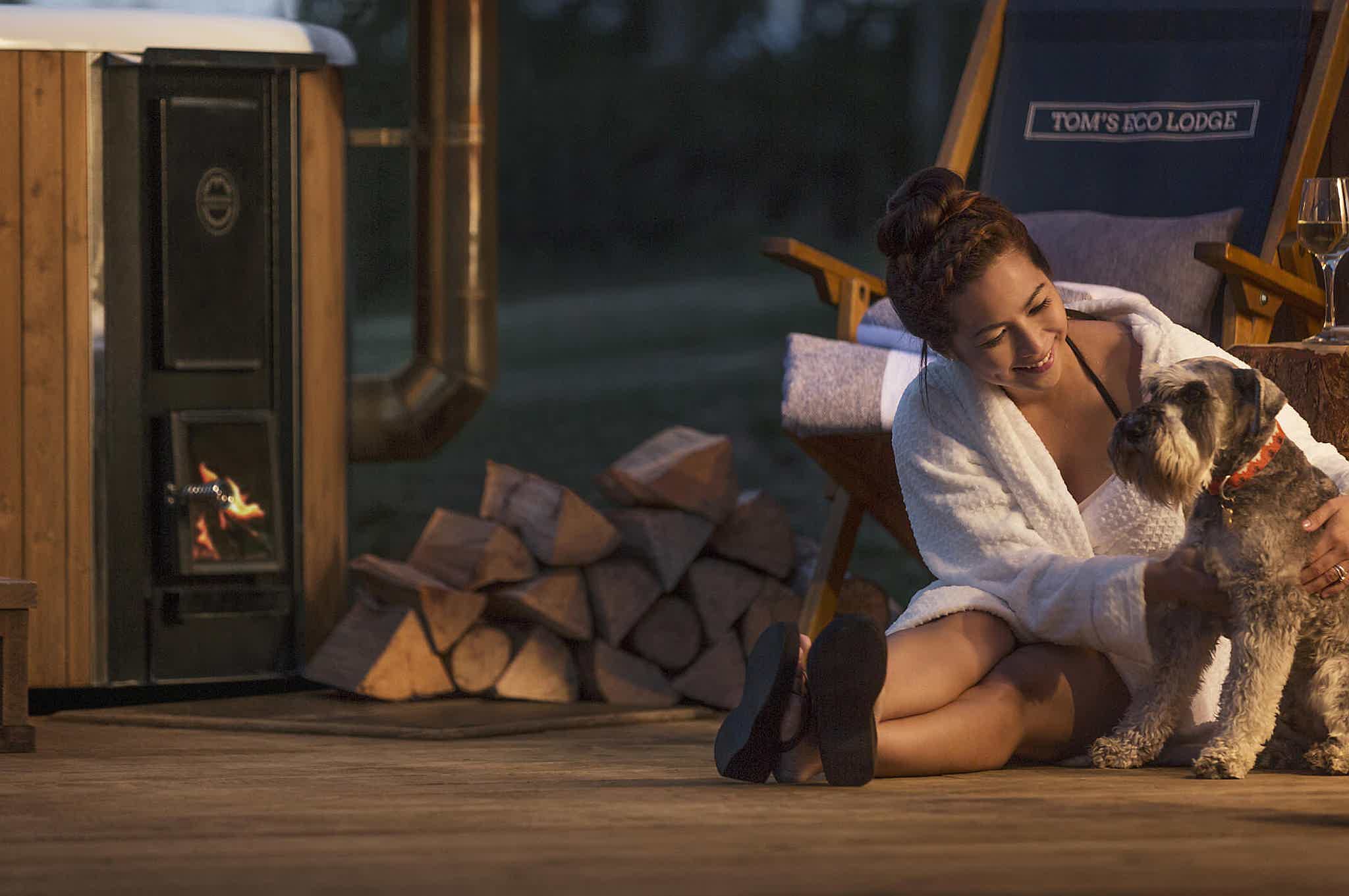 A serene moment with a woman in a white bathrobe and a Schnauzer by a wood-fired Skargards hot tub, logs, and fireplace.