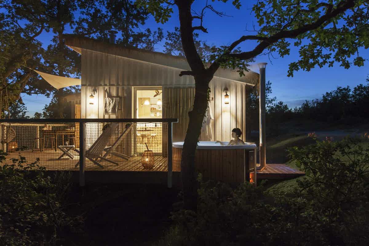 A cozy cabin with warm lights in a lush forest, offering a tranquil retreat with a wood-fired Skargards hot tub.