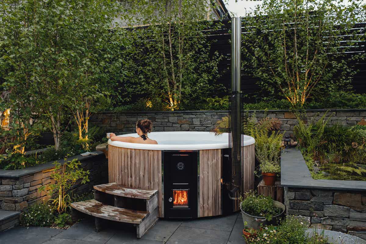 A cozy garden with a grey Skargards Regal 220 hot tub on a stone patio, stone wall, and warm lights. Victoria in tub.
