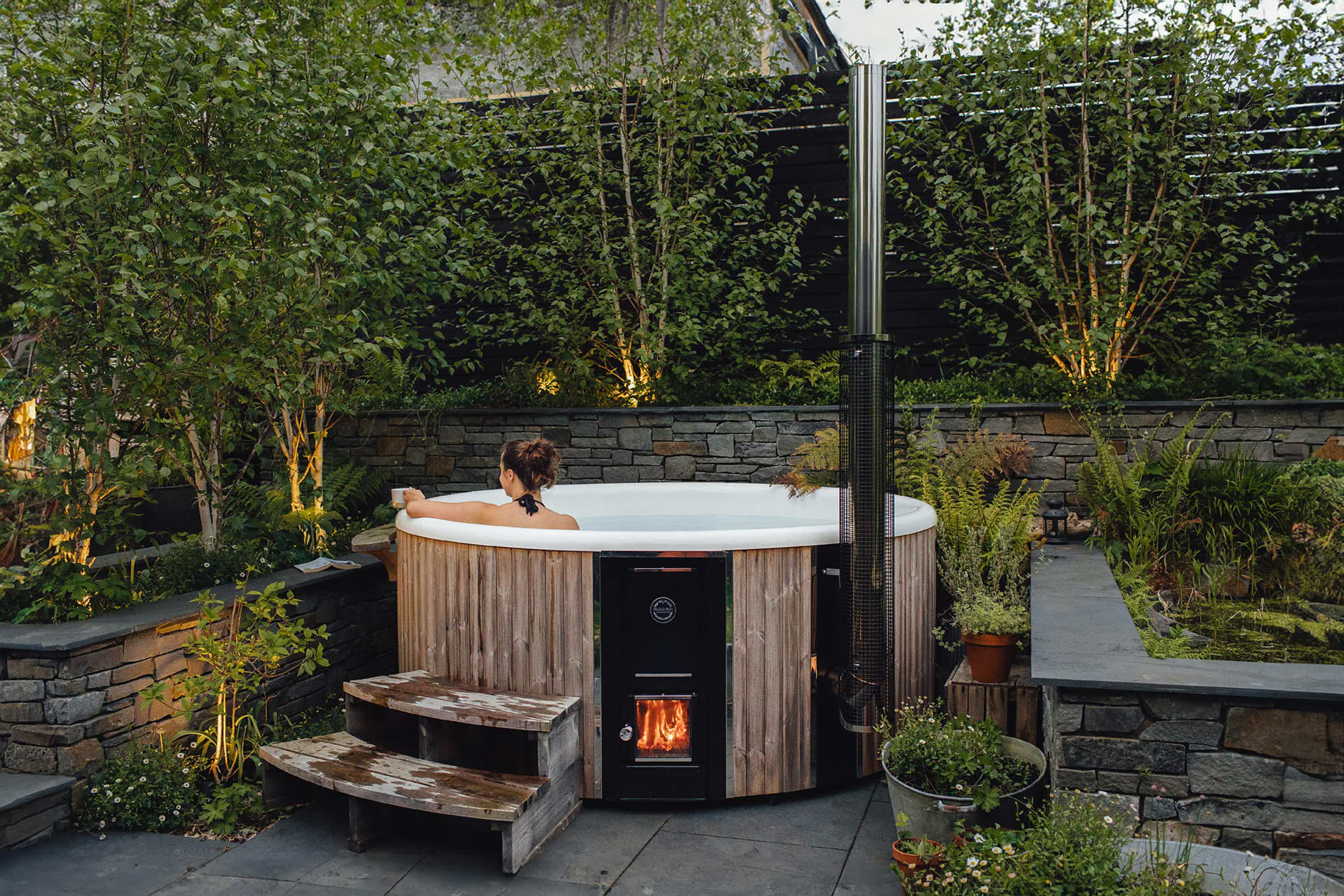 A cozy garden with a grey Skargards Regal 220 hot tub on a stone patio, stone wall, and warm lights. Victoria in tub.