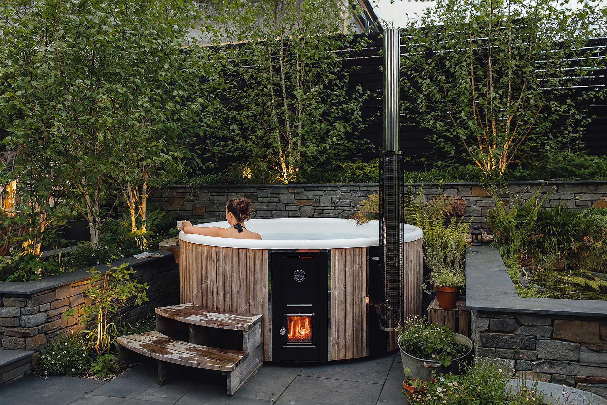 A cozy garden with a grey Skargards Regal 220 hot tub on a stone patio, stone wall, and warm lights. Victoria in tub.