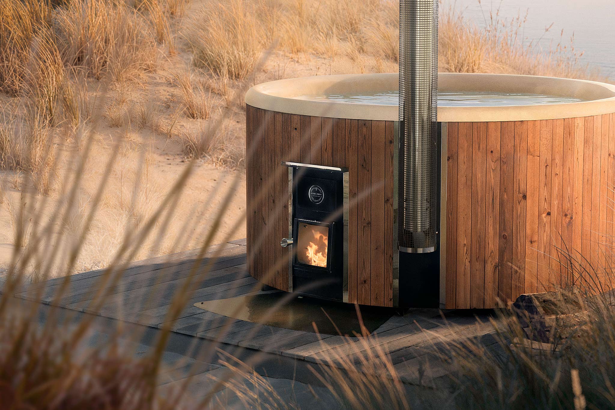 Modern wood-fired hot tub on black platform by beach, surrounded by grass, with light and glass door.