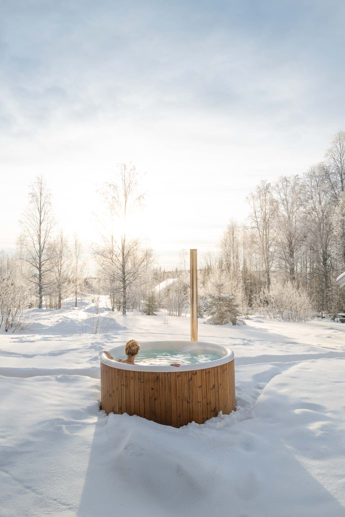 A wooden hot tub in a snowy landscape, surrounded by trees and a clear sky.