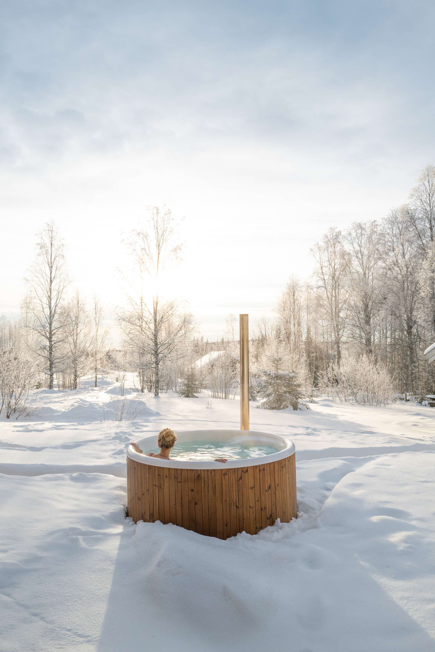 A wooden hot tub in a snowy landscape, surrounded by trees and a clear sky.