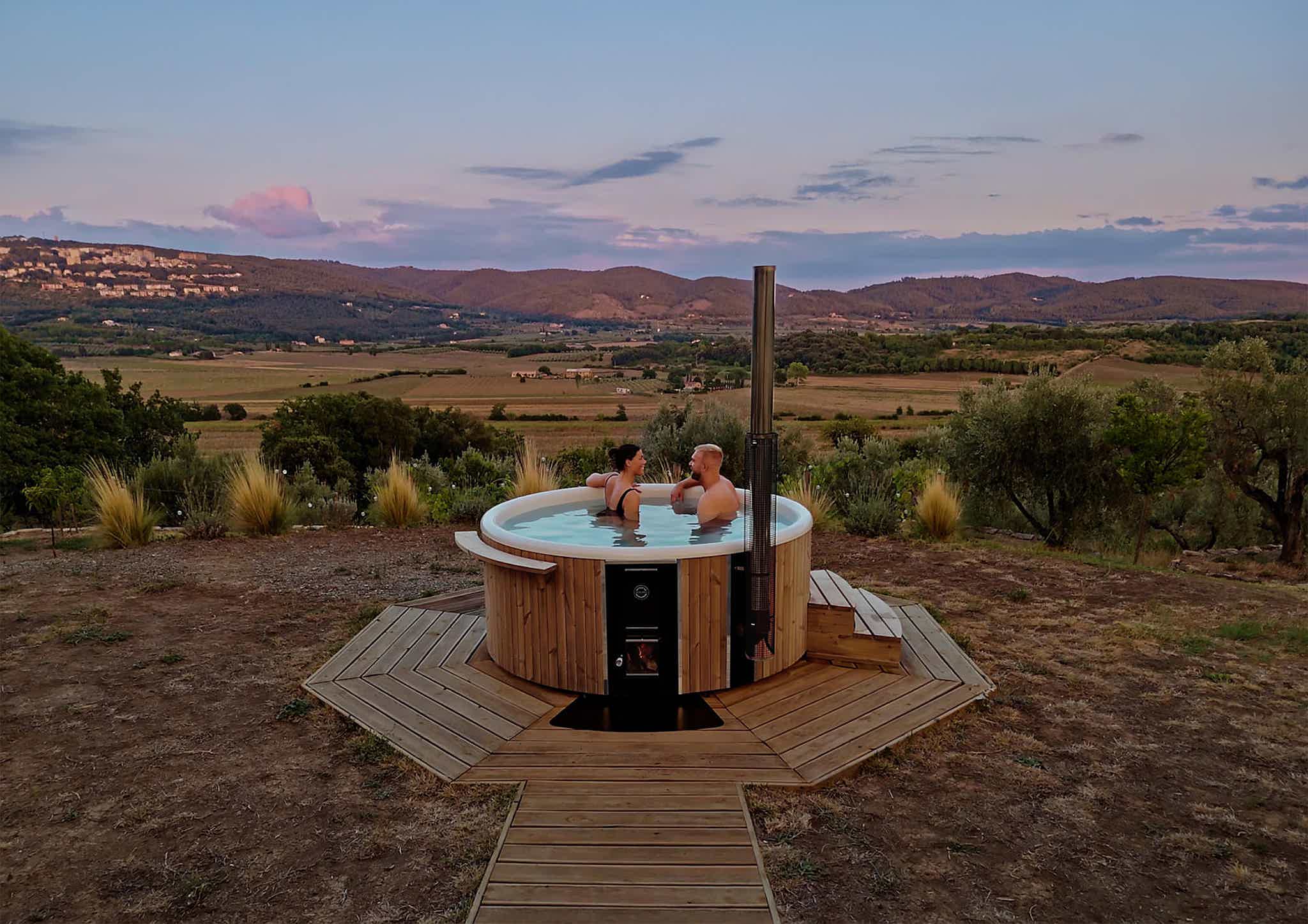 Two people in a Skargards Regal hot tub on a deck with scenic hills and valley at sunset in Tuscany.