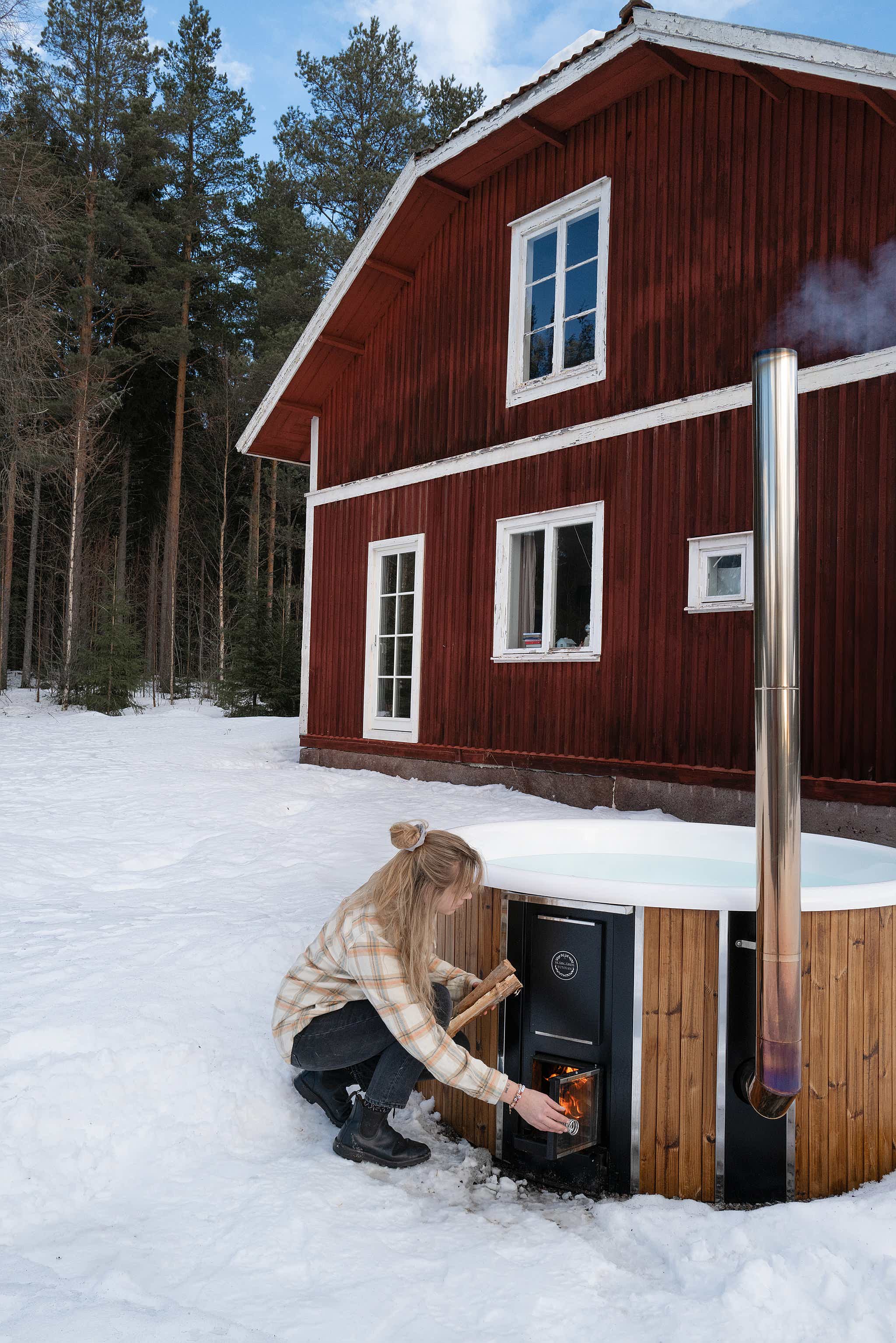 A woman cleans a wooden hot tub in a snowy forest, holding a water bottle. The tub has a chimney and window.