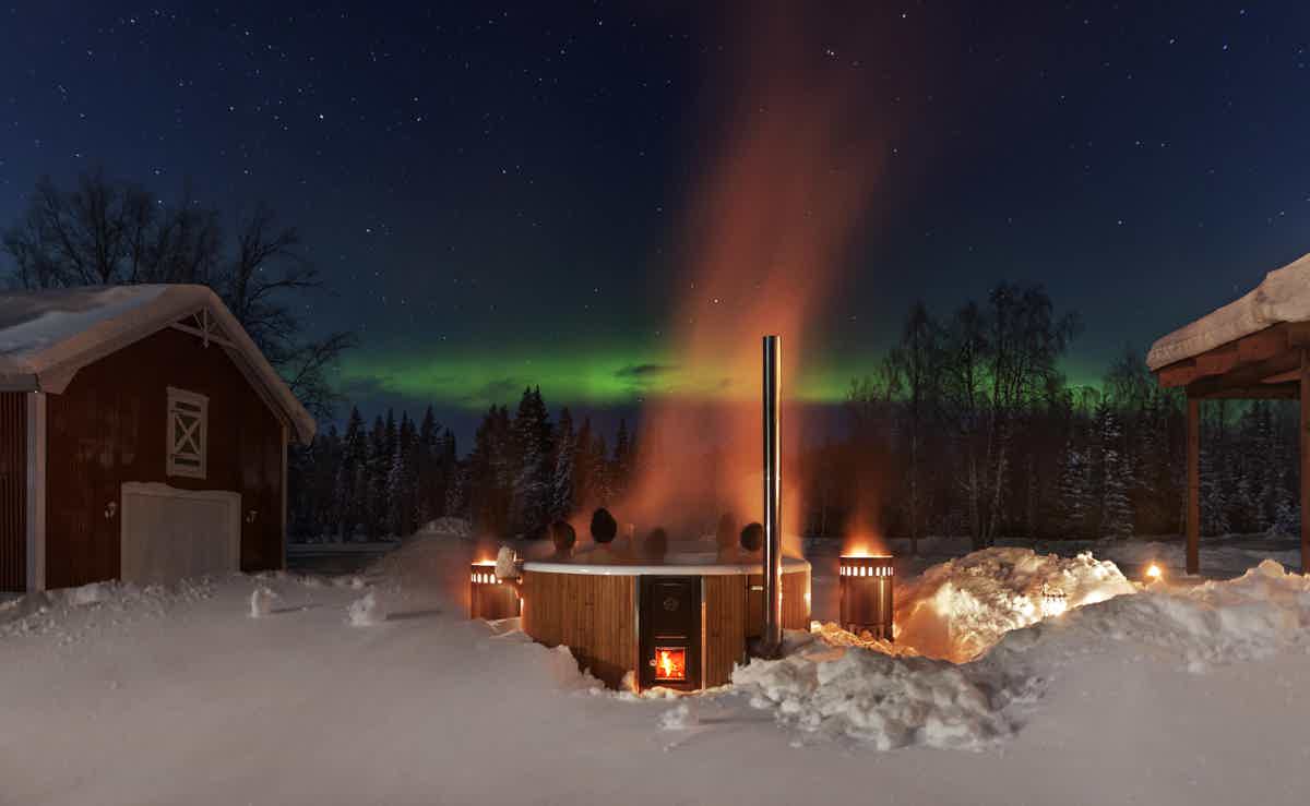 A cozy scene with a heated Skargards hot tub in front of snowy woods under the aurora borealis.