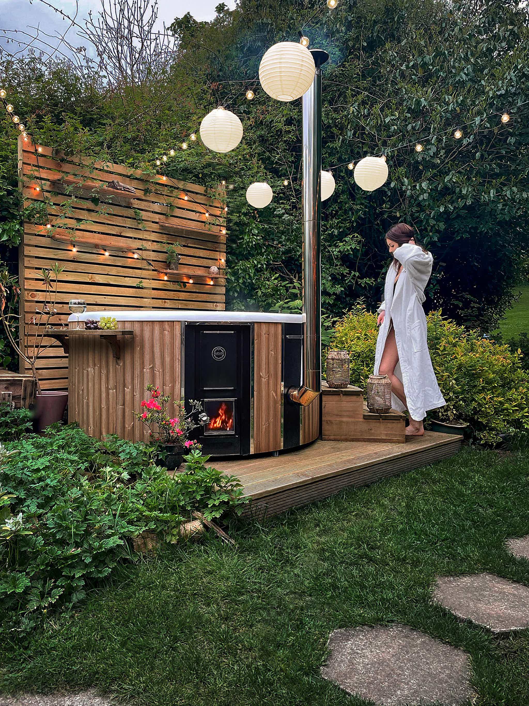 A woman in a white robe stands on a wooden deck near a Skargards Regal 190 hot tub with greenery and string lights above.