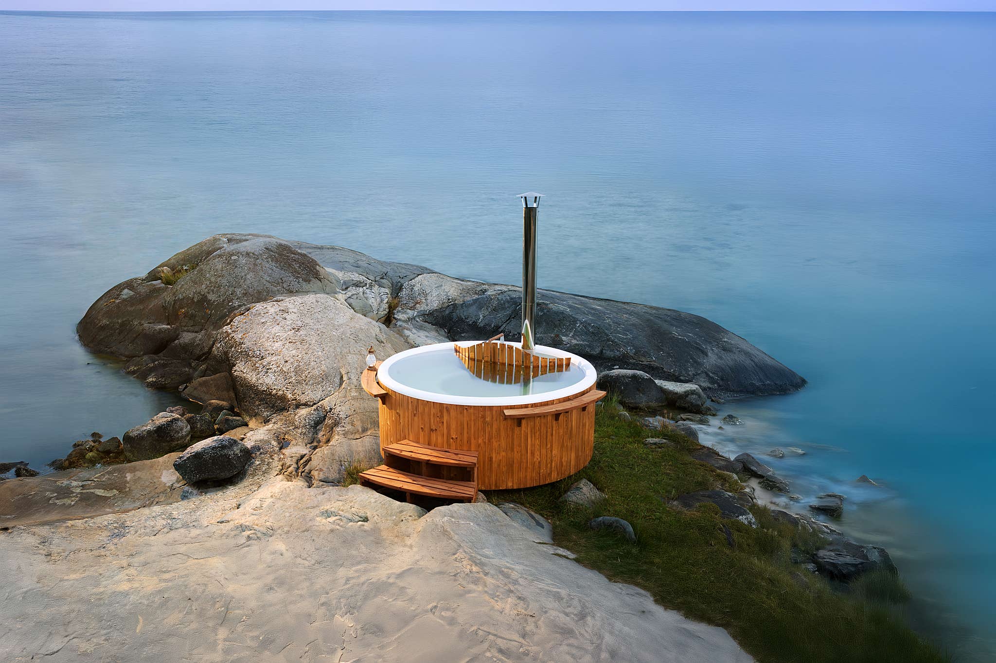 A wooden hot tub on a rocky shore with a metal pipe, serene blue water and horizon.