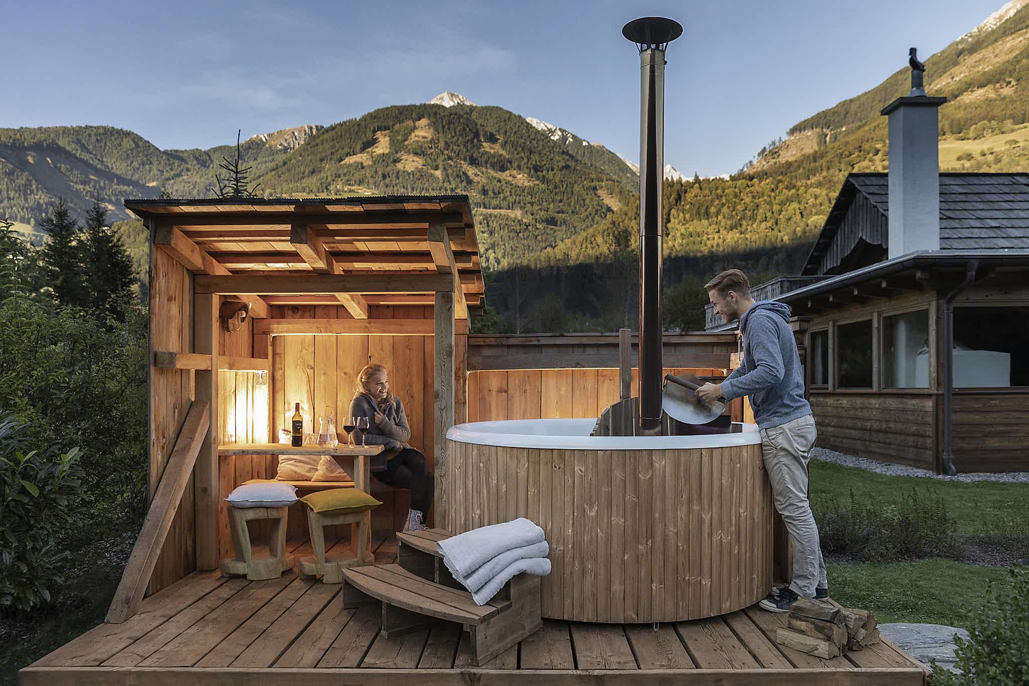A couple enjoys a chill day on a wooden deck with a Skargards Panel hot tub. The man heats the stove, mountains behind.