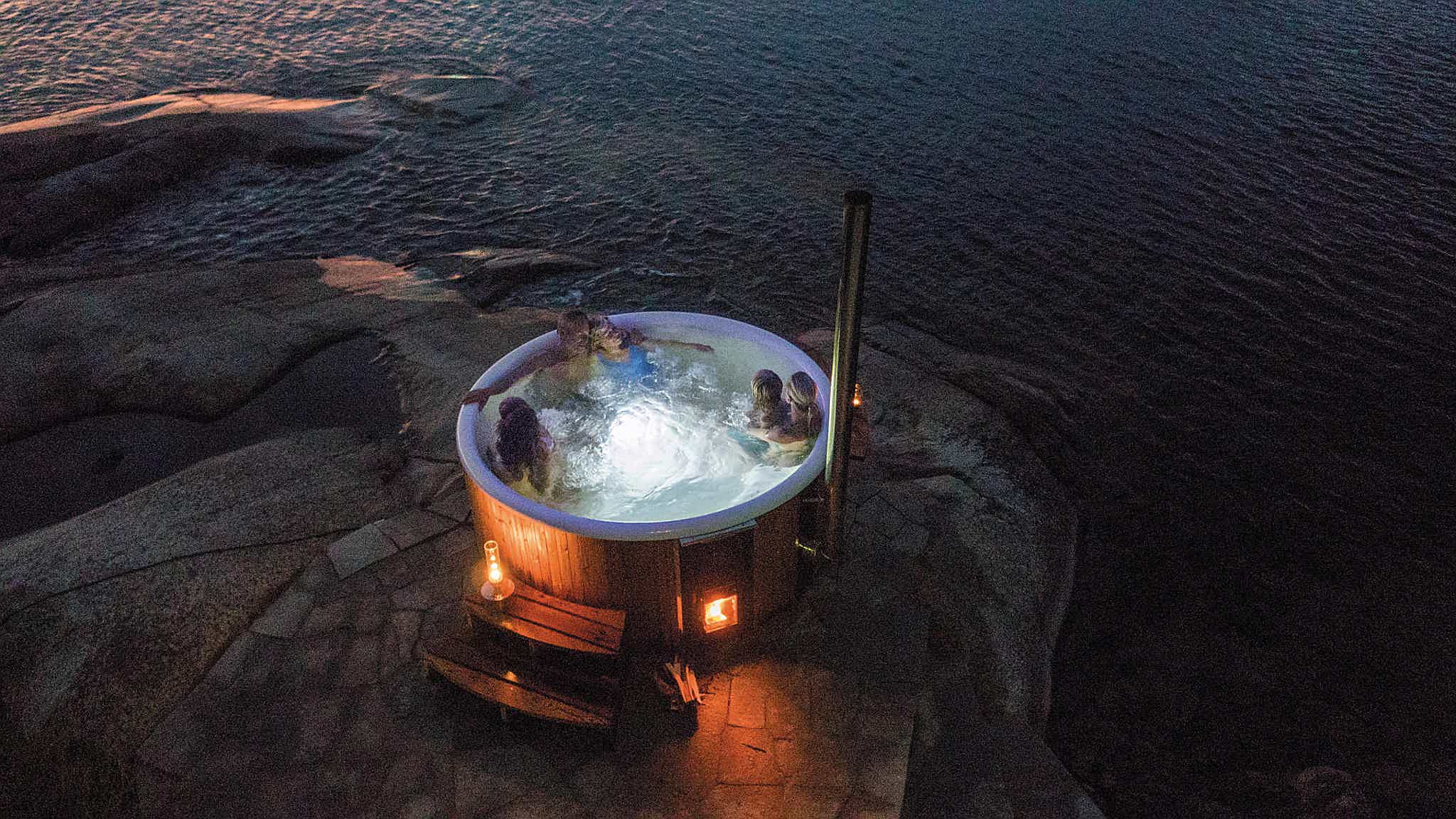 A wood-fired Skargards hot tub on a rocky shore, overlooking water. A family of six enjoys the tub, candles create serene atmosphere.