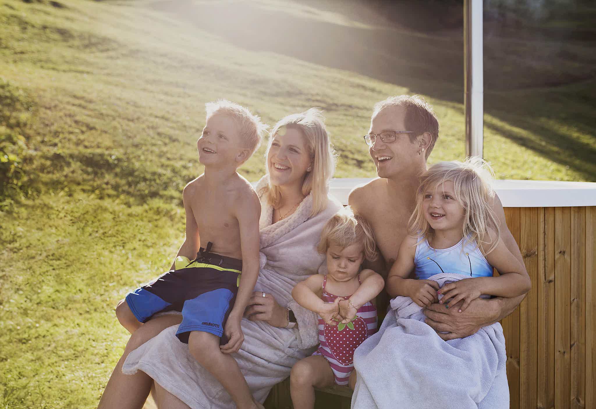 Family of five relaxing on the steps of a wooden Skargards Regal hot tub, with grassy hill backdrop.