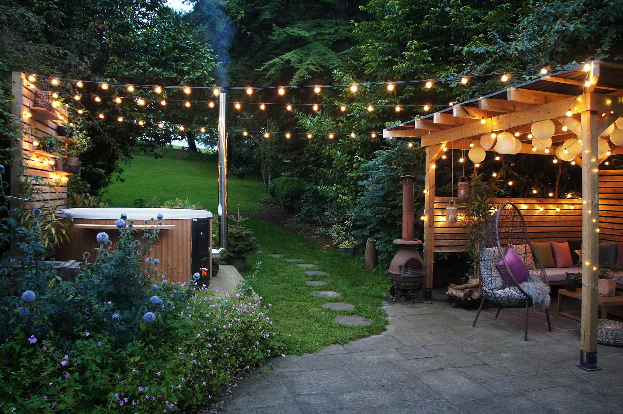 A peaceful backyard with a wooden gazebo, patio furniture, and lush greenery. A wood-fired Skargards hot tub sits on a wooden deck to the left.