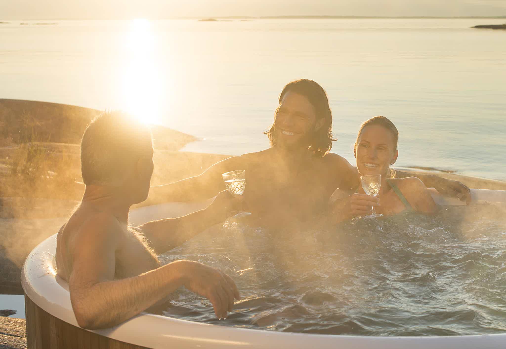 Family chatting in a Skargards Regal hot tub at sunset, surrounded by water and rocky shore. Bubble system is on.