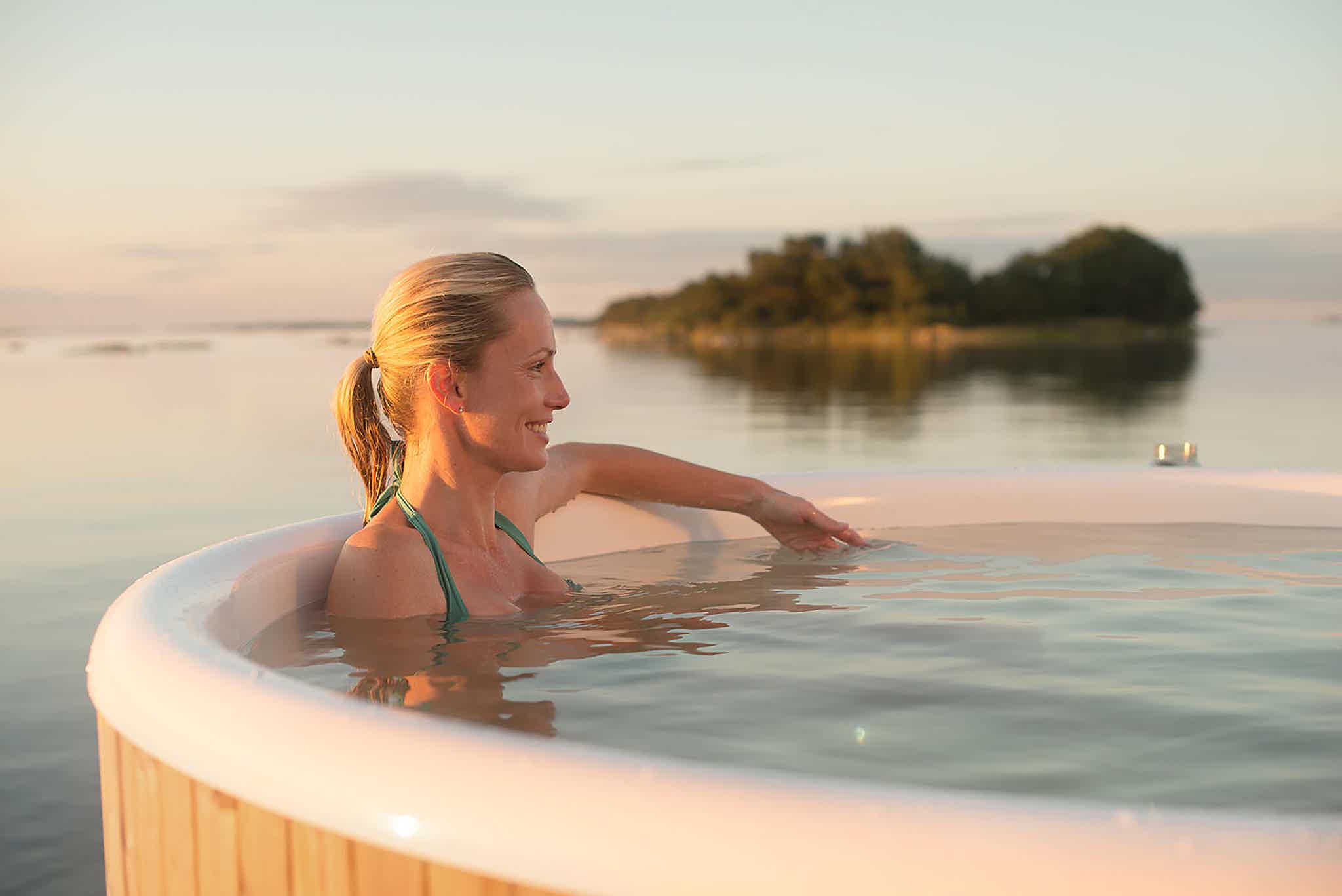 A woman relaxes in a Skargards Regal hot tub with a clear sky and distant island.