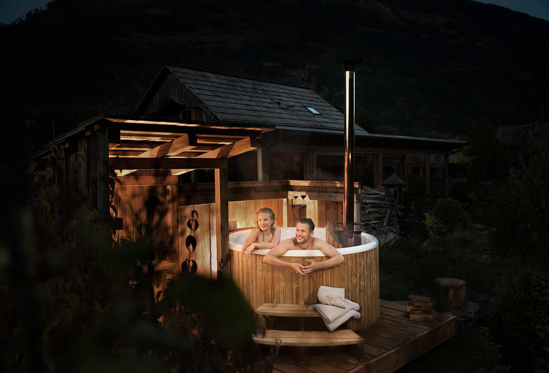 A couple relaxes in a Skargards Panel 190 hot tub on a wooden deck, surrounded by nature.