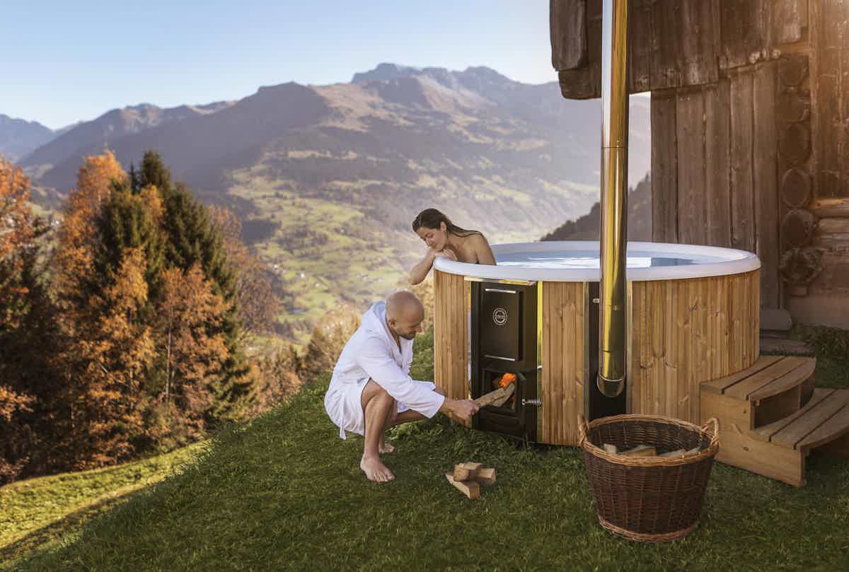 A man in a white bathrobe heats up a Skargards Regal 190 hot tub on a hillside, surrounded by mountains and a clear sky.