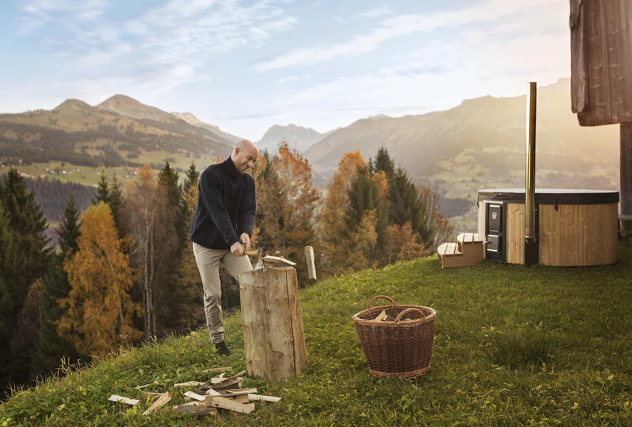 A man chopping wood on a hill with mountains, blue sky, and a Skargards Regal hot tub in the background.